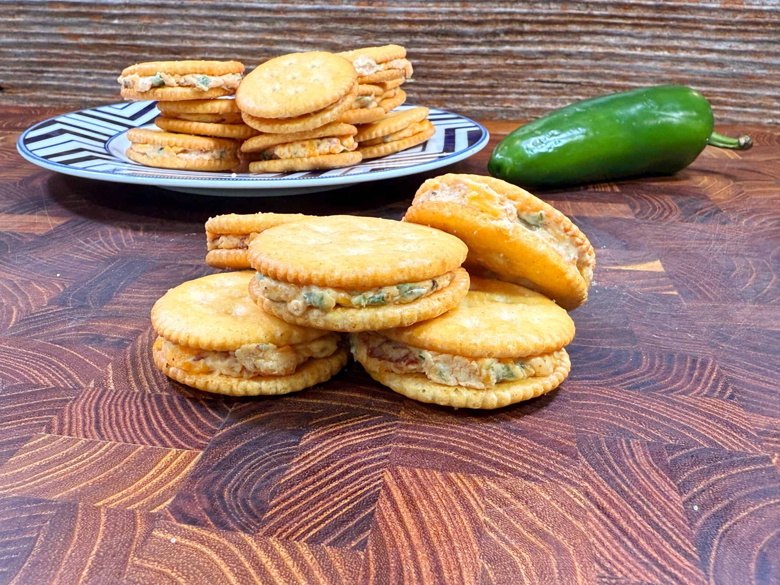 A stack of round jalapeno popper  crackers filled with a creamy mixture sits on a wooden surface, with a plate of similar crackers and a whole jalapeño pepper in the background.