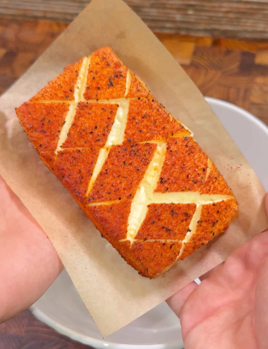 A person holds a rectangular, diagonally scored piece of golden, seasoned bread with a creamy filling, resting on a sheet of parchment paper.