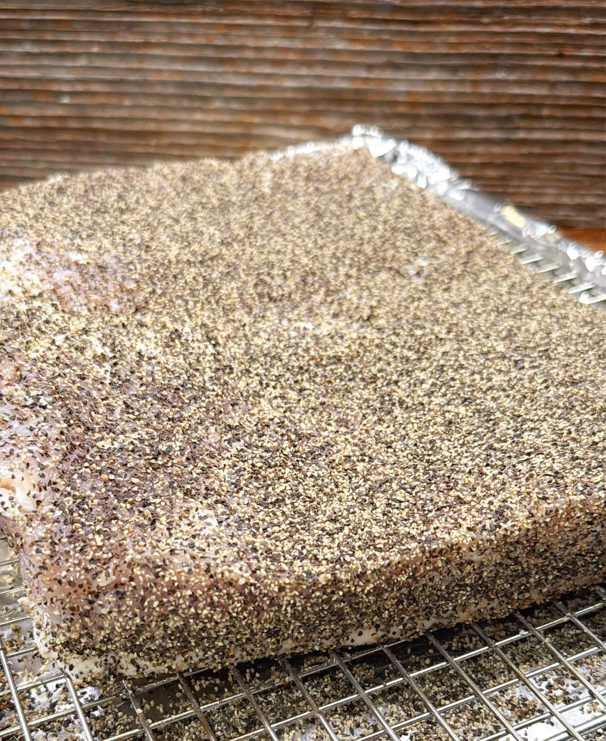 A slab of raw meat generously coated with cracked black pepper sits on a wire rack over a foil-lined tray, ready for curing or smoking. The background features a wooden surface.