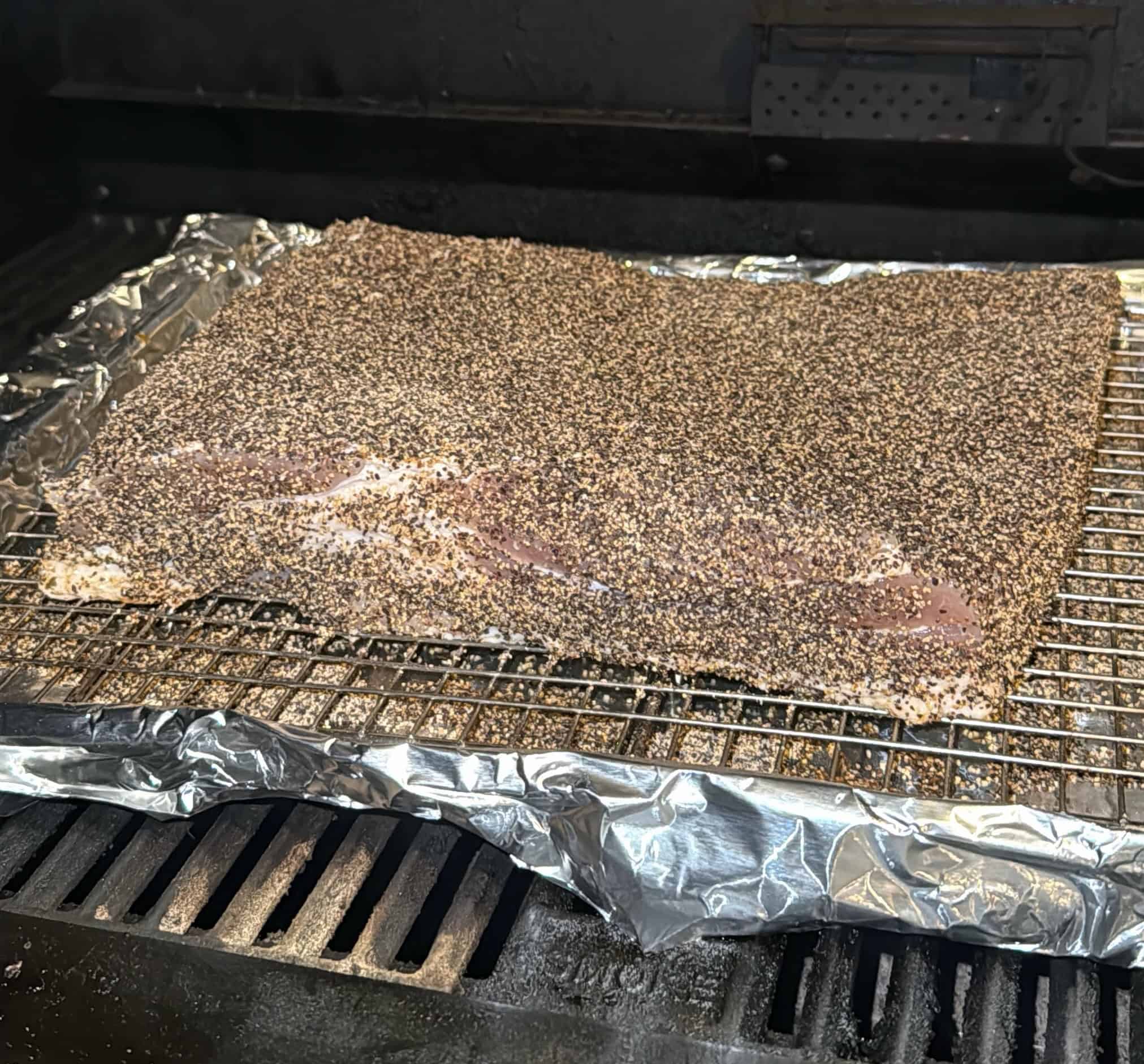 A large, uncooked brisket coated heavily with pepper and spices sits on a wire rack covered with foil inside a smoker or grill, ready to be cooked.