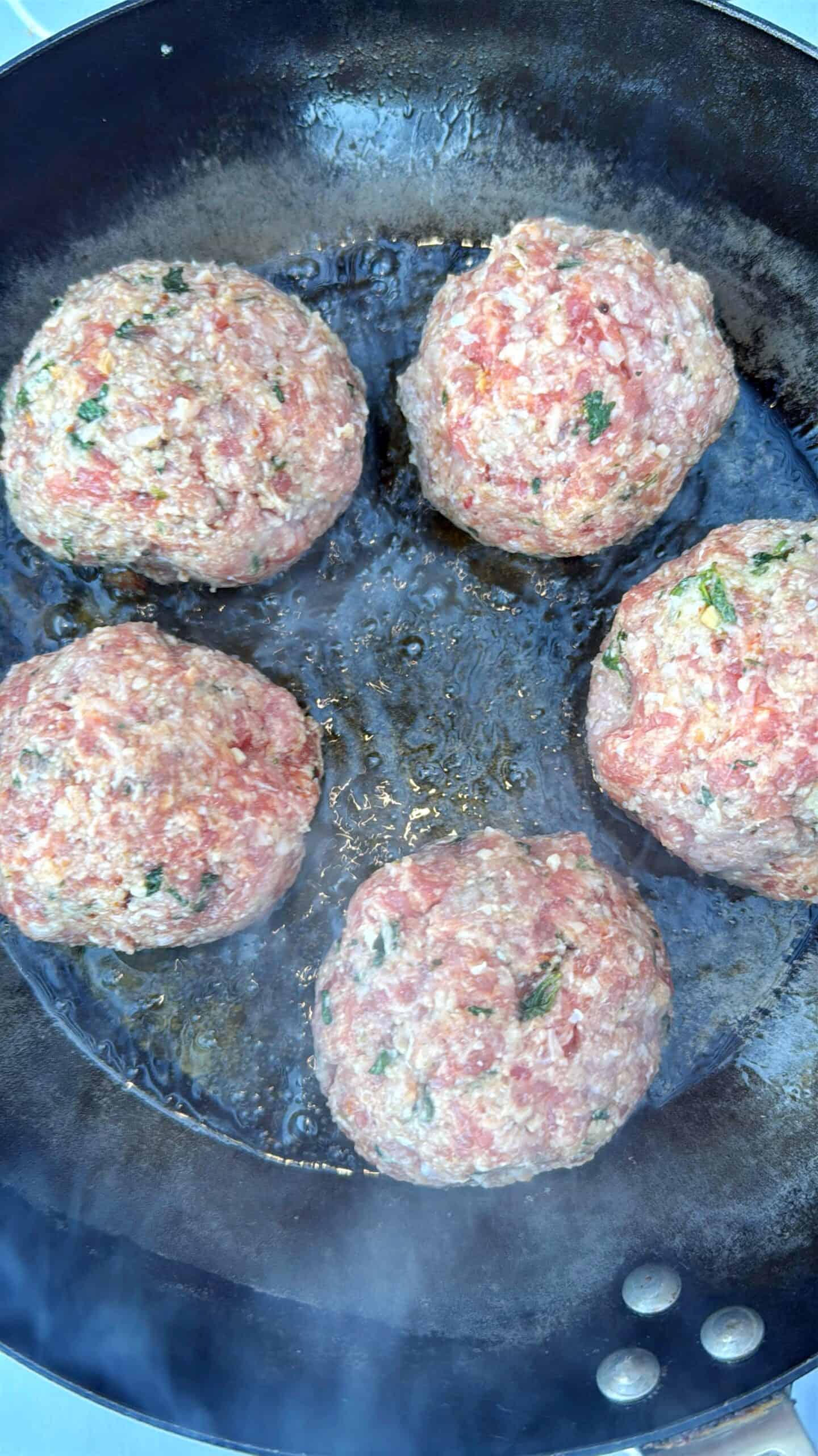 Five raw meatballs with herbs are sizzling in a black frying pan, beginning to cook. Steam is rising from the pan, and the meatballs are spaced evenly across the surface.