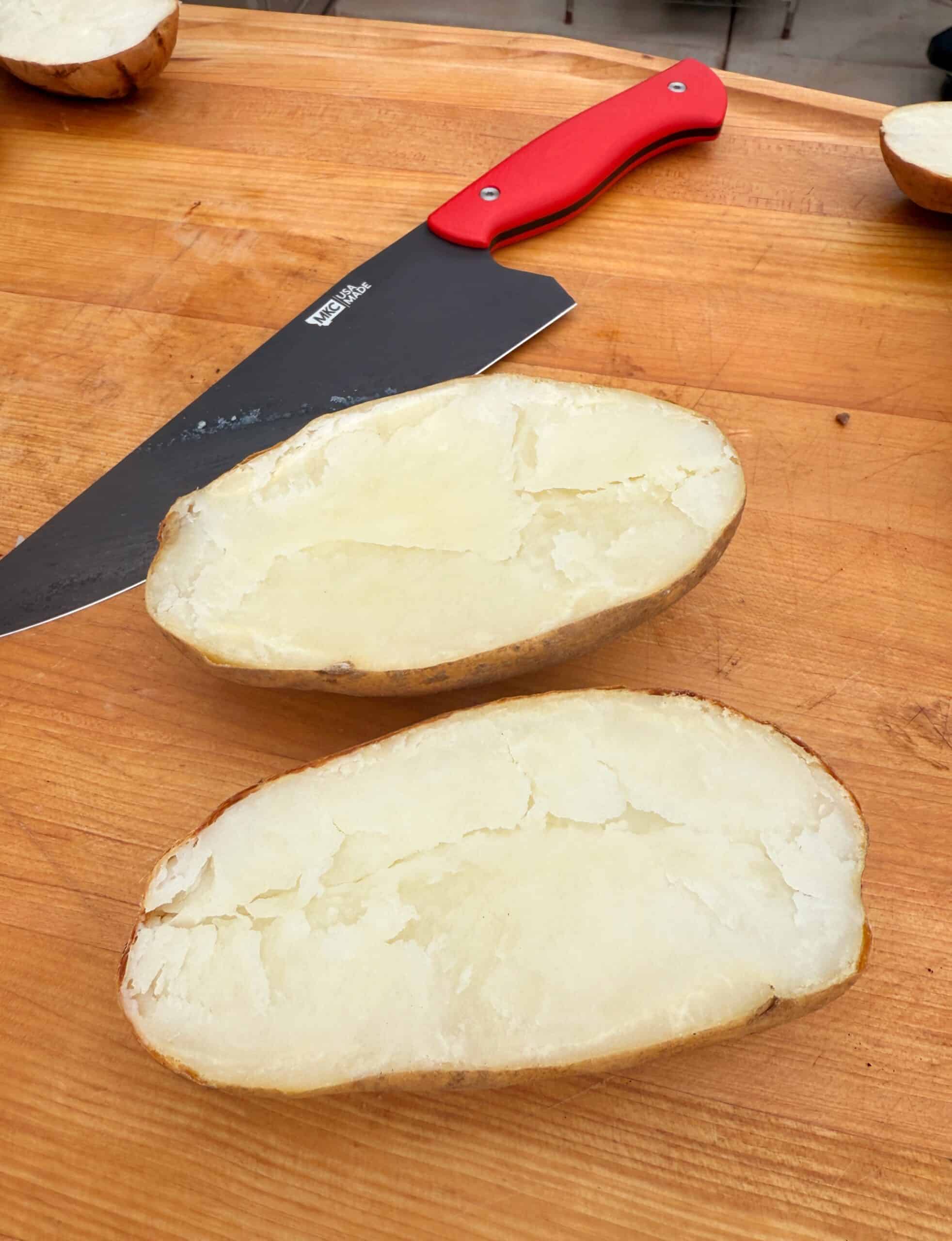 A large baked potato sliced in half on a wooden surface, with a red-handled knife lying nearby. The inside of the potato looks soft and flaky.