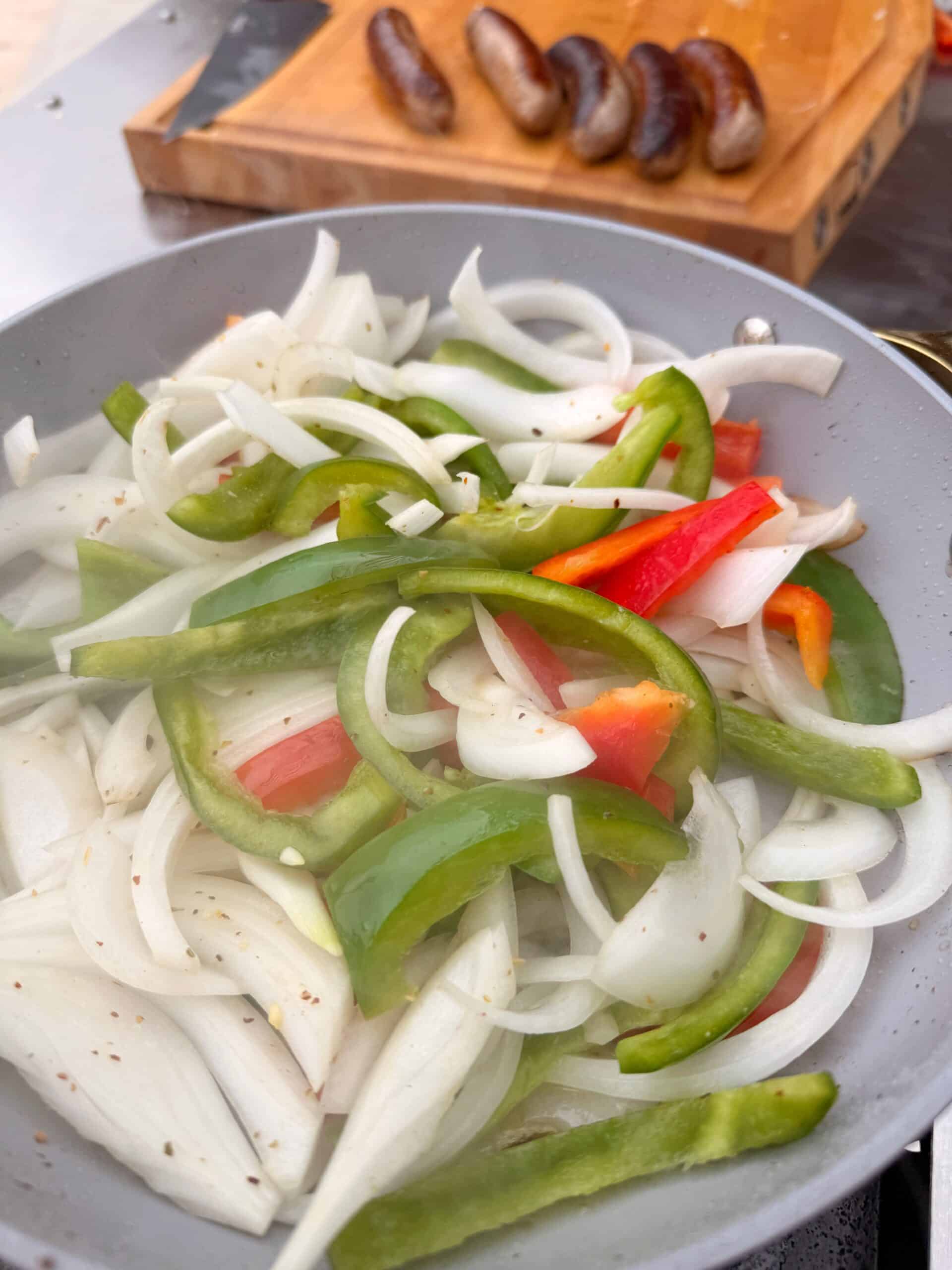 Sliced onions and bell peppers cooking in a pan, with sliced grilled sausages on a wooden cutting board in the background.
