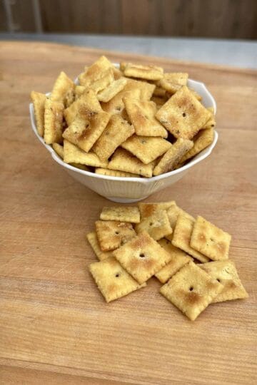 A white bowl filled with square, golden-brown crackers sits on a wooden surface, with several crackers scattered in front of the bowl.