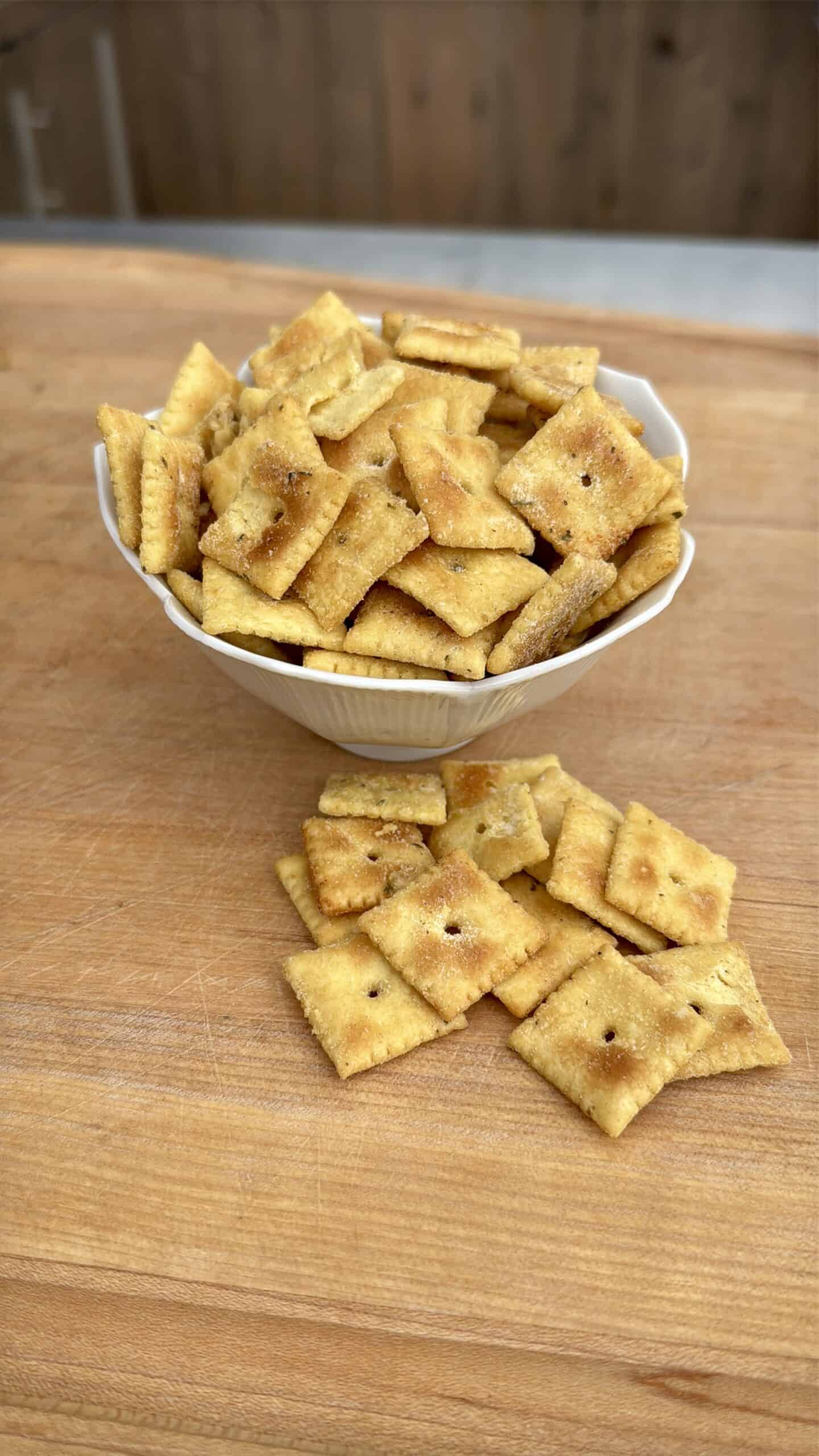 A white bowl filled with square, golden-brown crackers sits on a wooden surface, with several crackers scattered in front of the bowl.