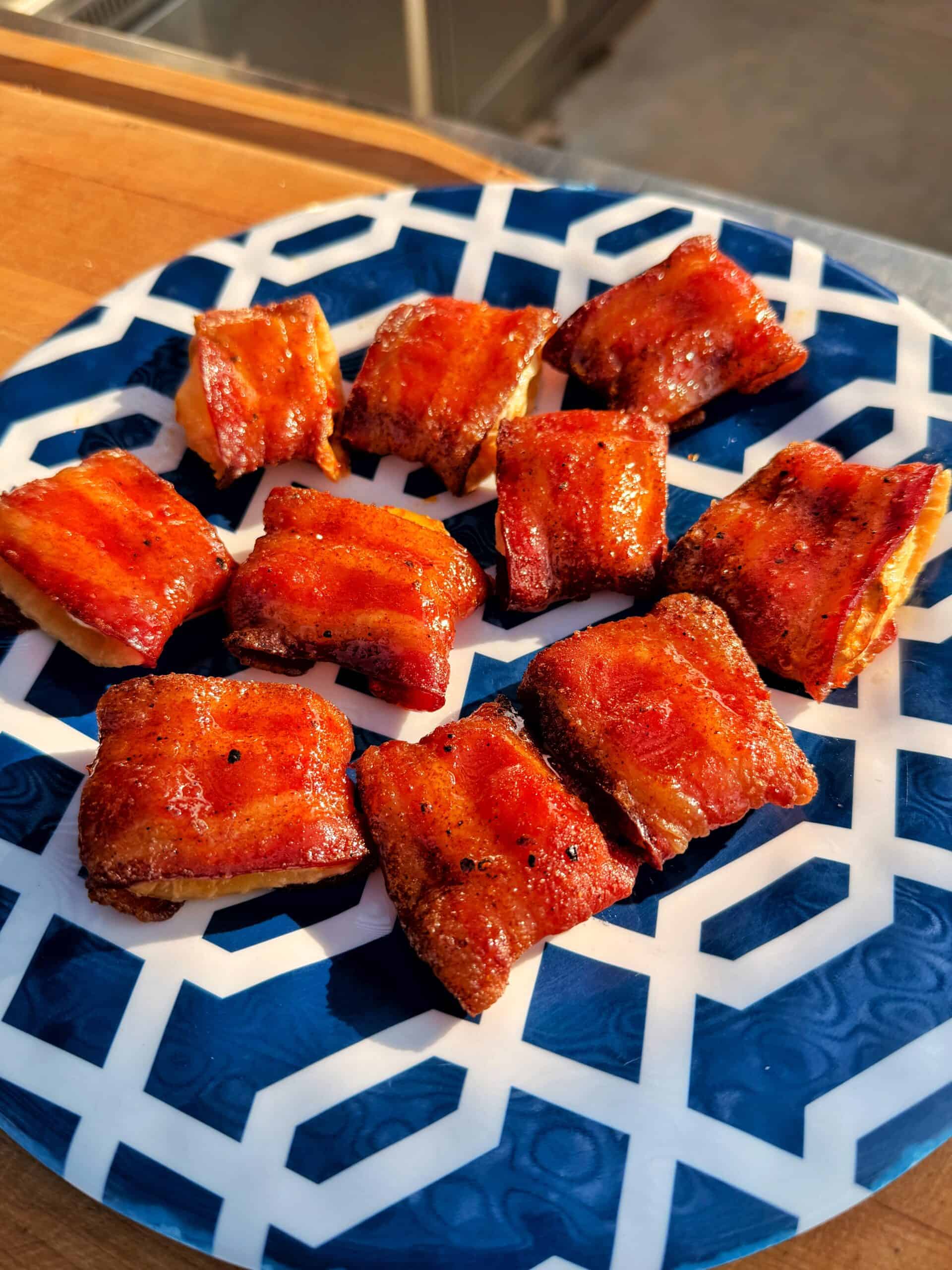 A blue and white patterned plate with several bite-sized pieces of bacon-wrapped appetizers, golden and crispy, arranged in rows and glistening in natural sunlight.
