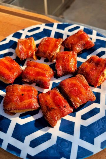 A blue and white geometric-patterned plate with several pieces of crispy, glazed bacon-wrapped bites arranged neatly in sunlight.
