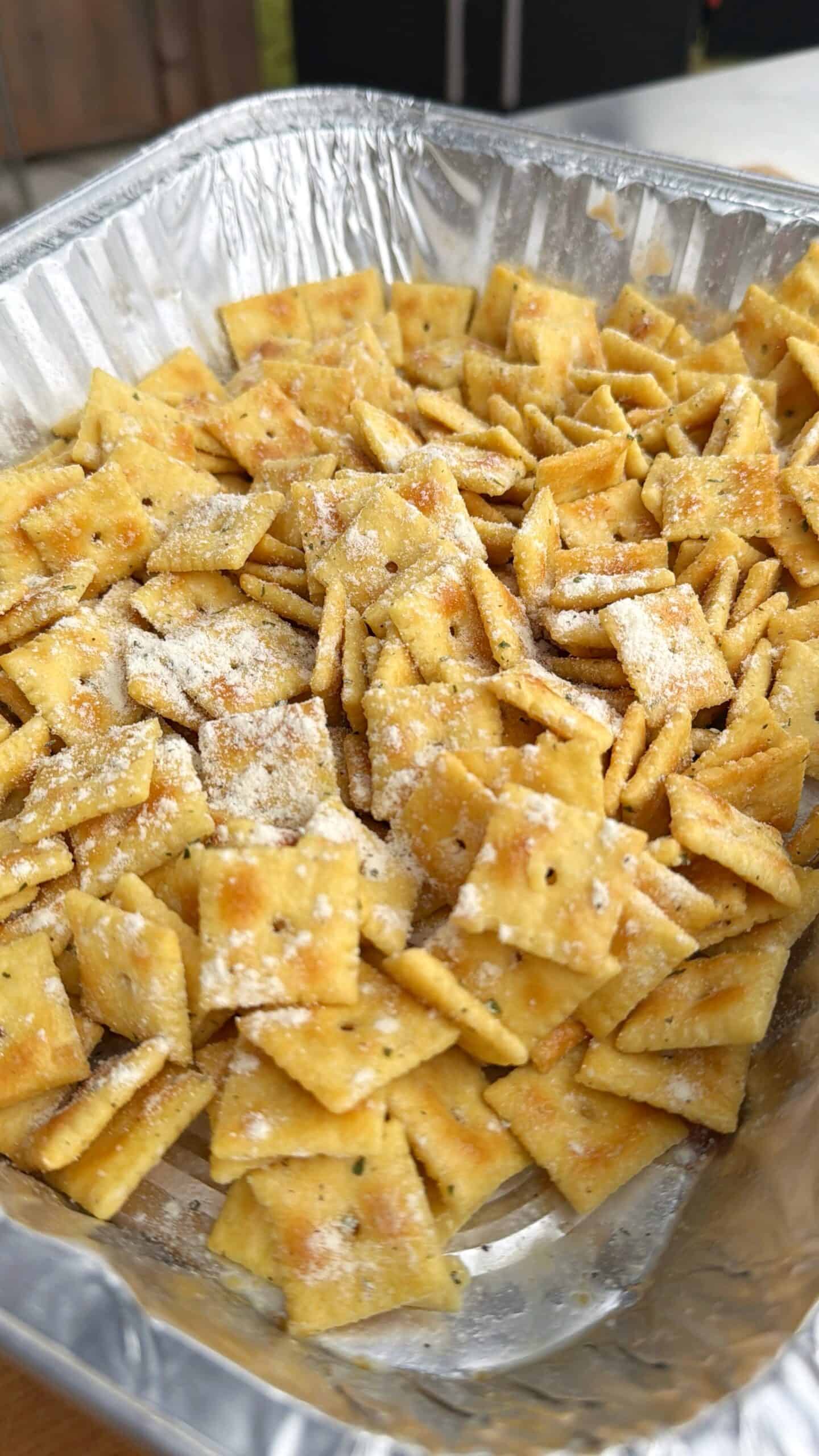 A close-up of a foil tray filled with seasoned square crackers, like Cheez Its, sprinkled with grated cheese or zesty jalapeno seasoning powder.