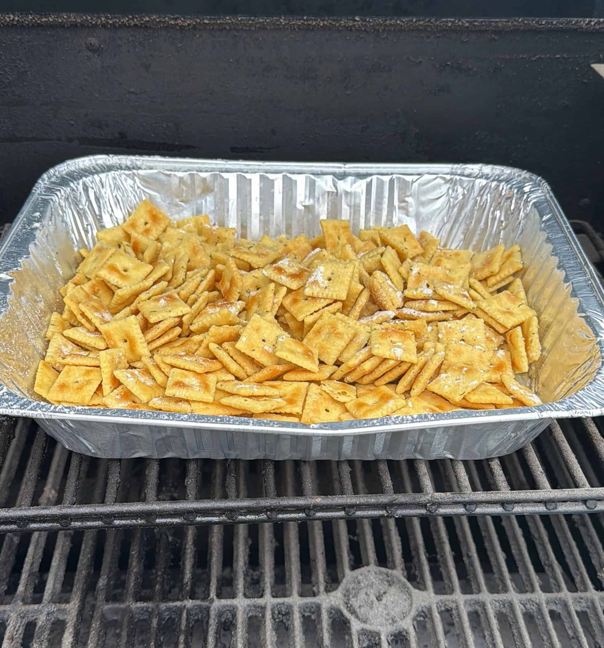 A disposable aluminum tray filled with Cheez Its, sprinkled with ranch seasoning, sits on a grill grate inside a barbecue smoker.