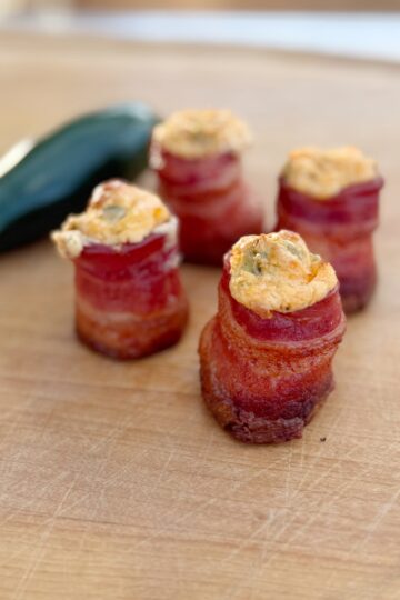 Four bacon-wrapped appetizers filled with a creamy, orange-colored mixture are arranged on a wooden surface. A whole green jalapeño pepper lies in the background.