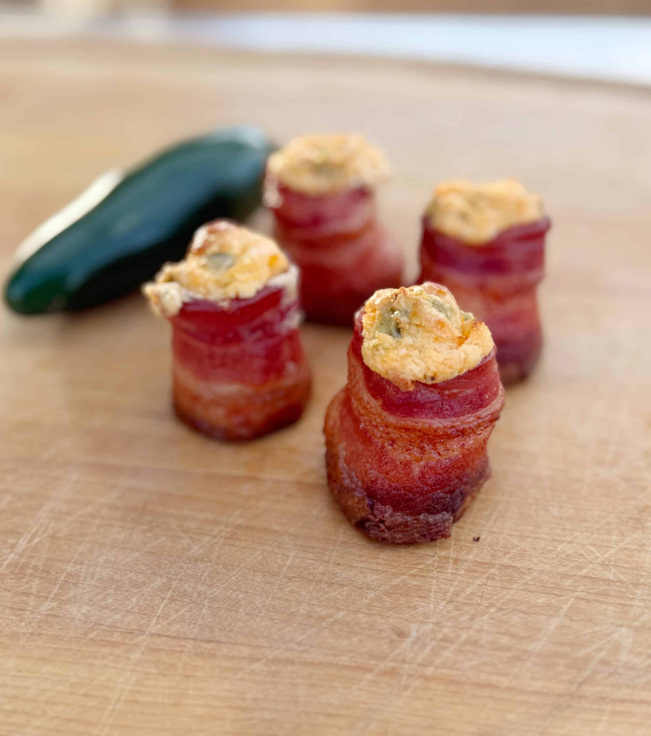 Four bacon-wrapped appetizers filled with a creamy, orange-colored mixture are arranged on a wooden surface. A whole green jalapeño pepper lies in the background.