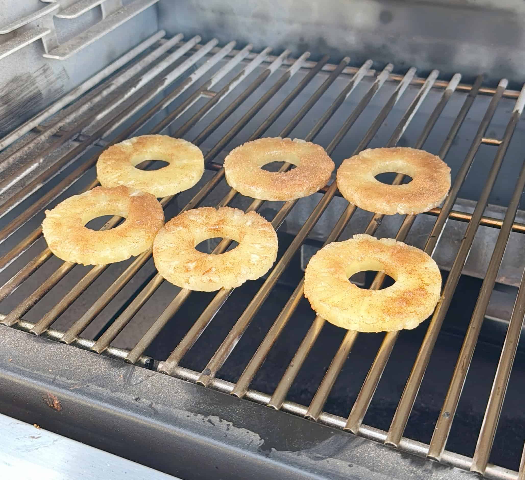 Six pineapple rings coated with a seasoning are being grilled on a barbecue, arranged in two rows on the metal grates. The grill is open and clean with no other food items present.