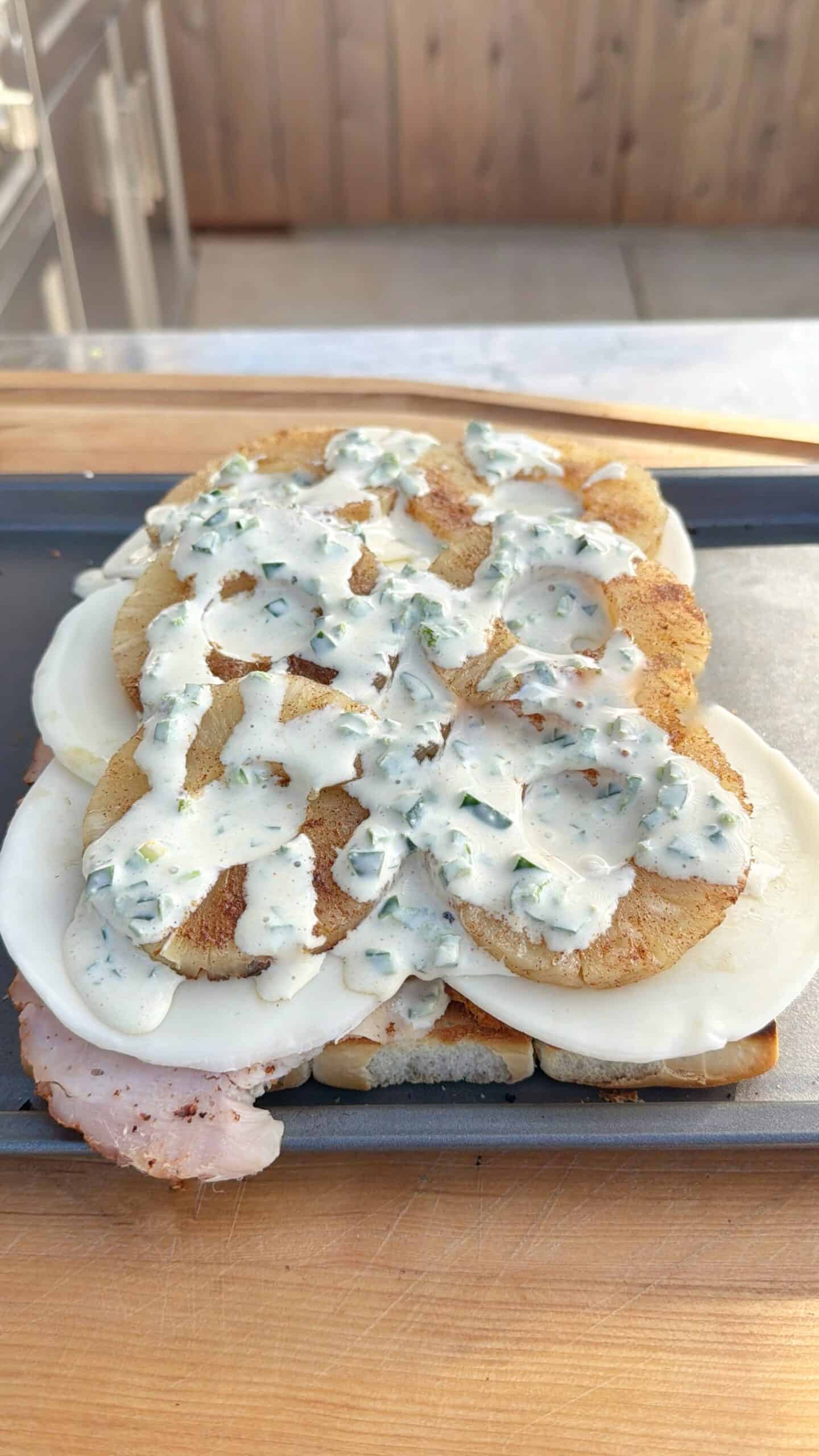 A sandwich being prepared with layers of grilled meat, sliced cheese, and fried potato rounds, topped with a creamy herb sauce on a tray.