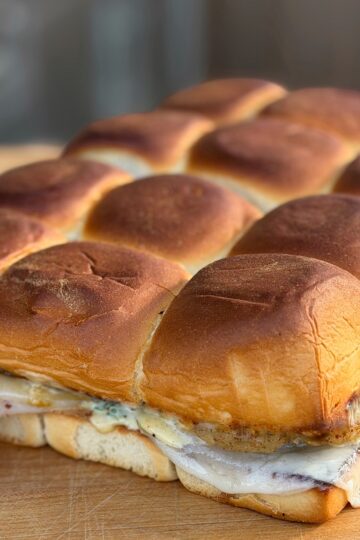 A close-up of a tray of slider sandwiches with golden brown, fluffy buns, melted cheese, and layers of deli meat and condiments, all sitting on a wooden surface.