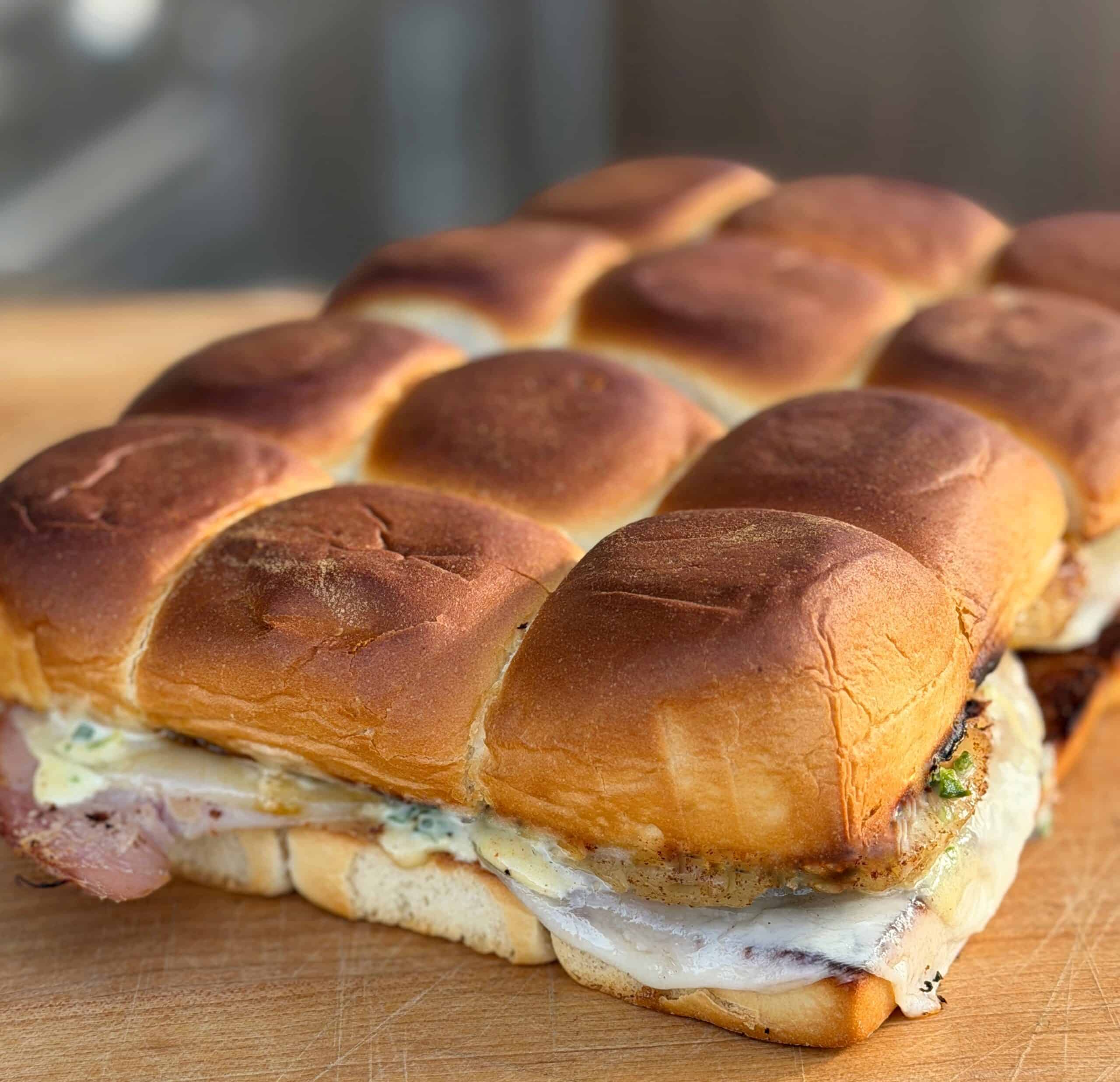 A close-up of a tray of easy Hawaiian sliders with golden brown, fluffy buns, melted cheese, and layers of deli meat and condiments, all sitting on a wooden surface.