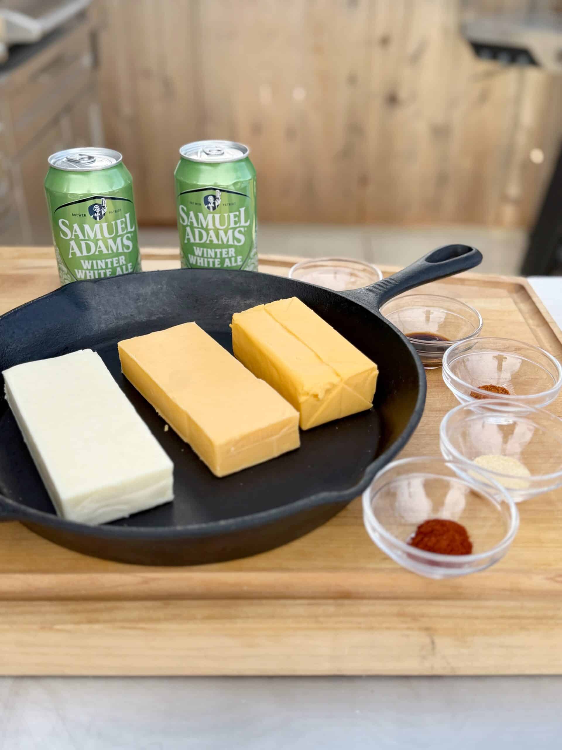 A cast iron skillet with white and yellow smoked cheese blocks sits on a wooden board. Surrounding it are clear bowls with spices and two cans of Samuel Adams Winter White Ale—perfect for your next smoked beer cheese recipe.