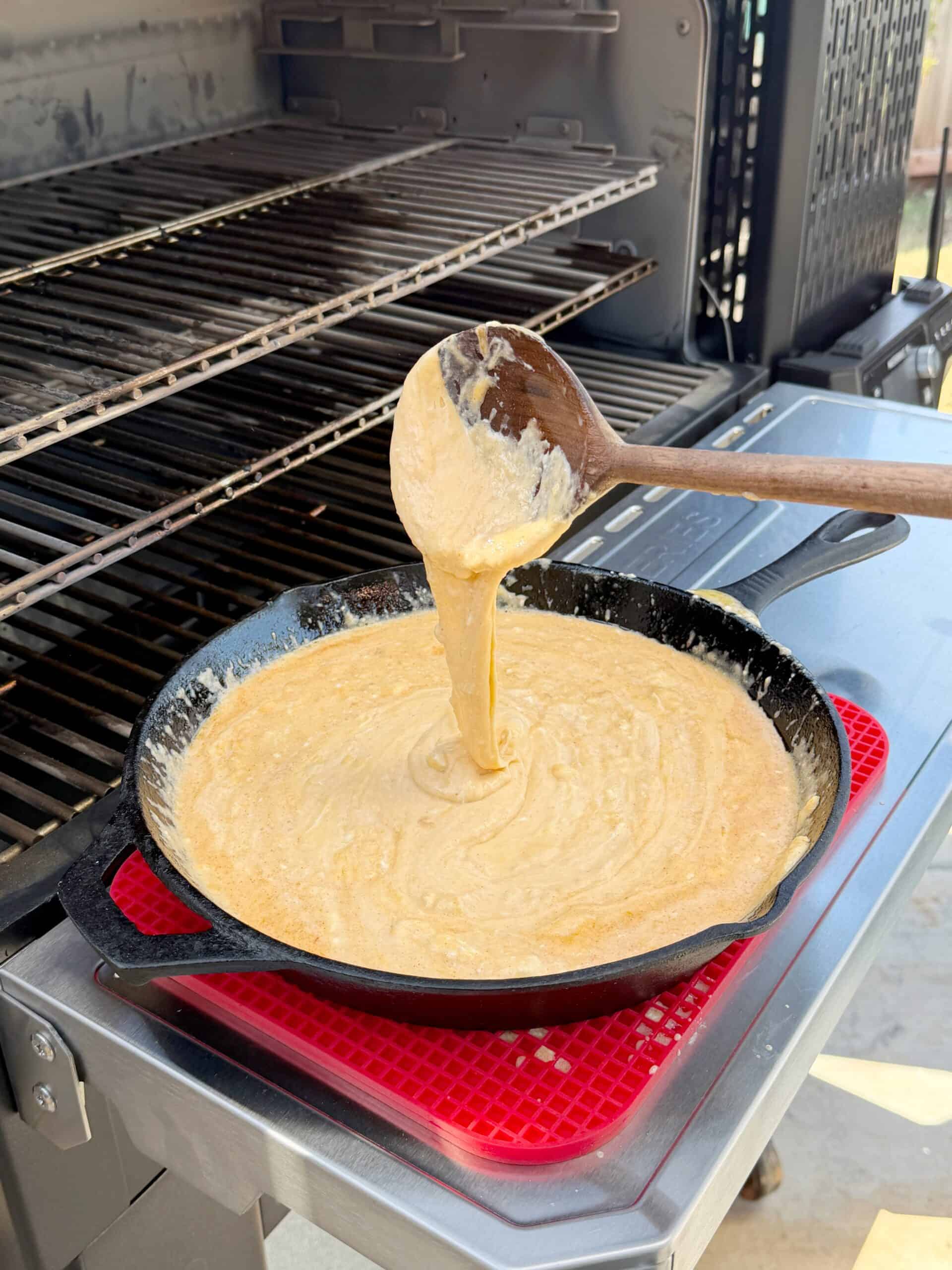 A wooden spoon lifts thick cornbread batter mixed with smoked beer cheese from a cast iron skillet sitting on an outdoor grill, with metal grates visible in the background.