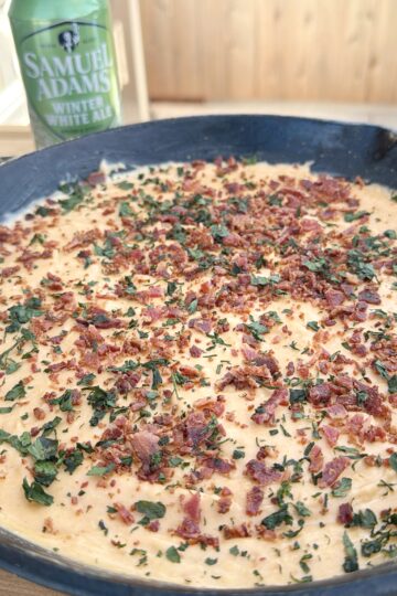 A close-up of a creamy smoked beer cheese dip, topped with chopped herbs and bacon bits in a dark dish, with a can of Samuel Adams Winter White Ale in the background on a wooden surface.