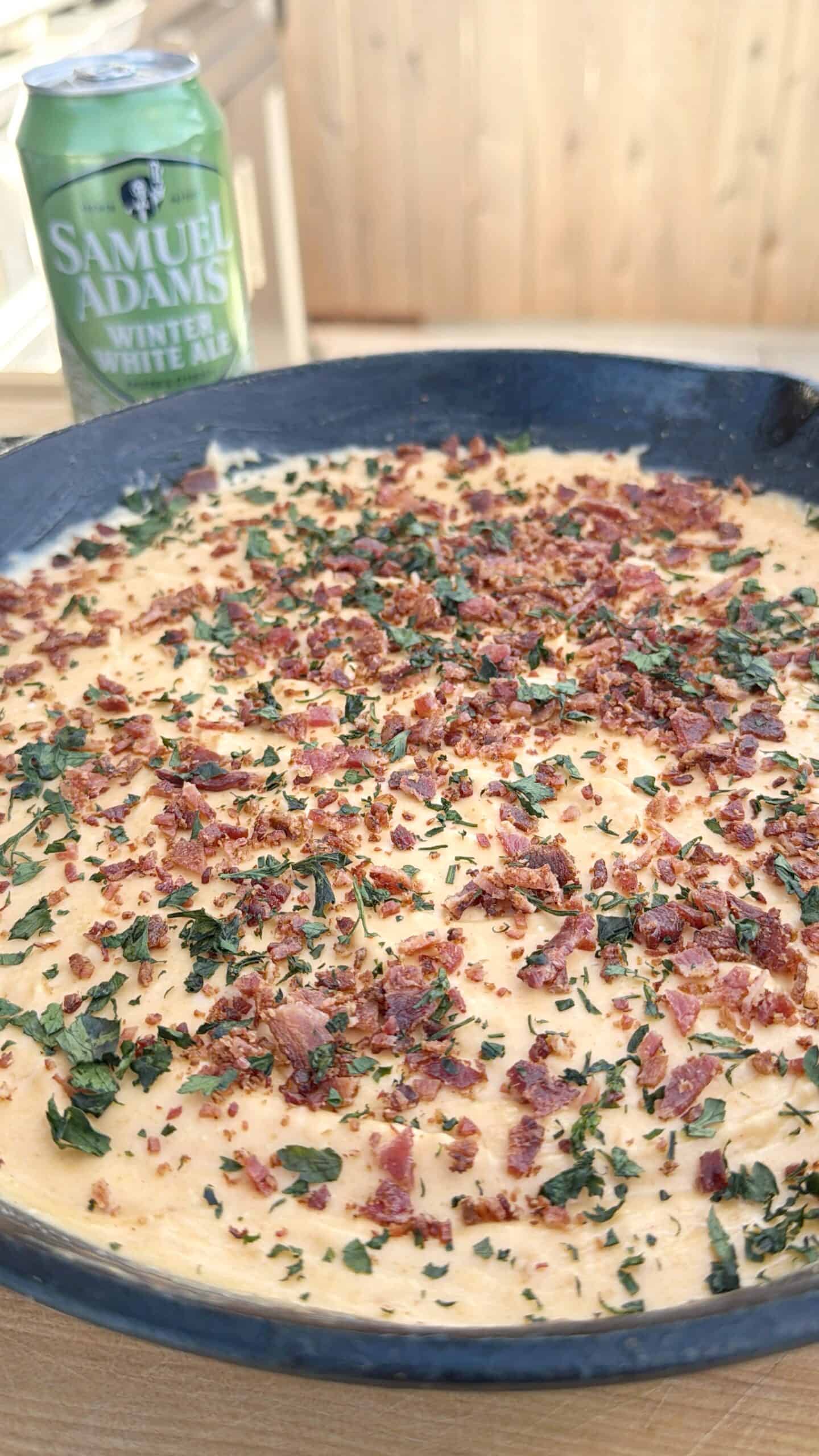 A close-up of a creamy smoked beer cheese dip, topped with chopped herbs and bacon bits in a dark dish, with a can of Samuel Adams Winter White Ale in the background on a wooden surface.