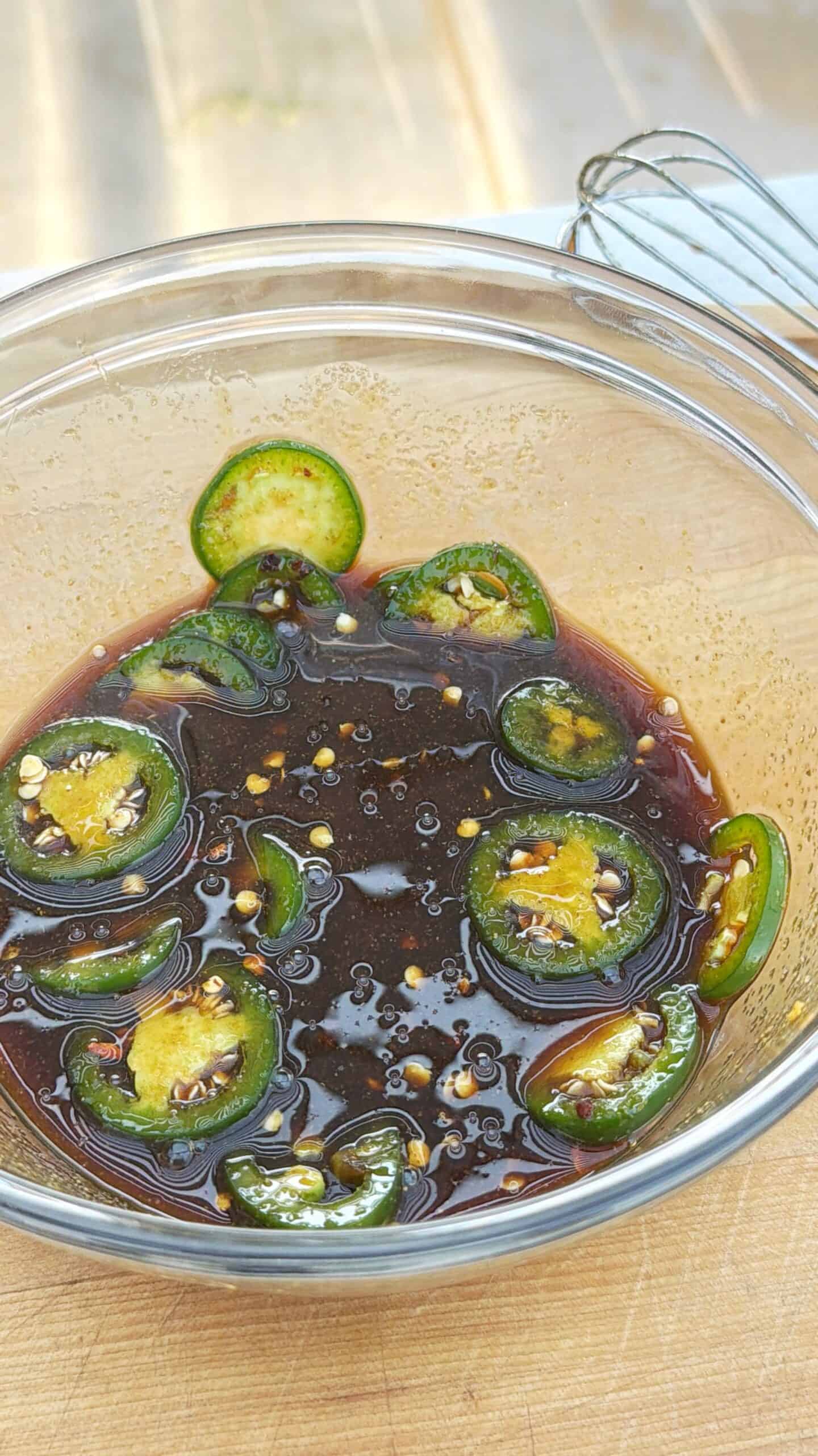 A glass bowl filled with a dark sauce, garnished with slices of fresh green jalapeño peppers and visible chili seeds, sits on a wooden surface with a metal whisk in the background.