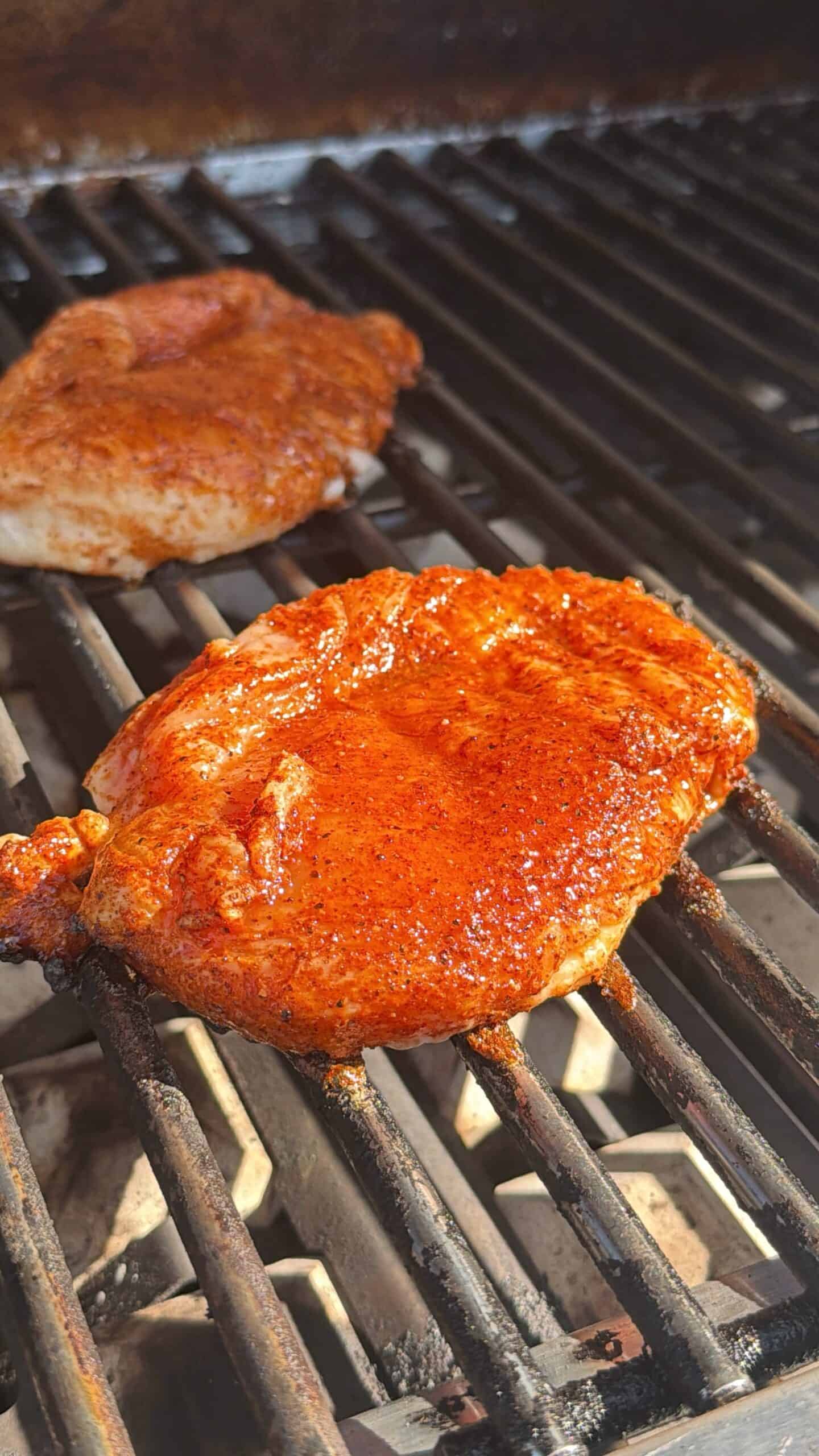Two pieces of seasoned meat are grilling on a barbecue, with one piece in the foreground covered in a reddish spice rub and the other piece in the background. Sunlight highlights the texture and seasoning.