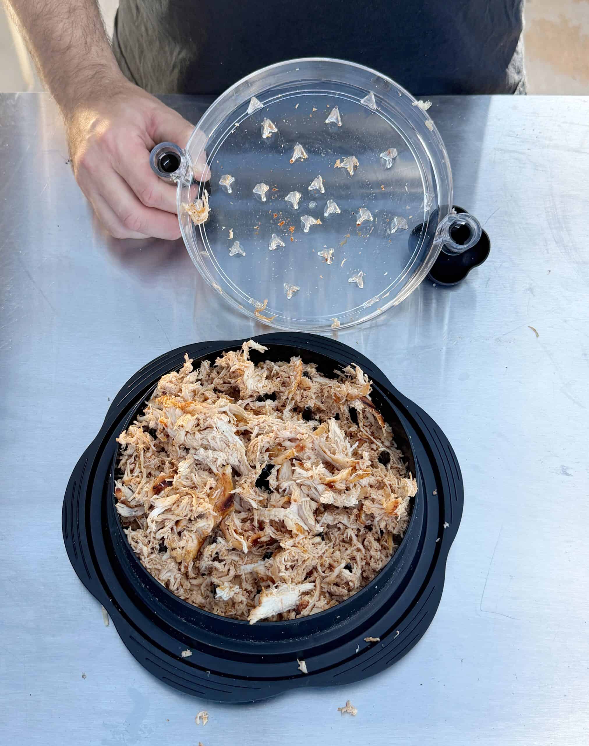 A person is lifting the lid off a black container filled with shredded cooked meat, placed on a stainless steel surface. Some meat pieces are stuck to the underside of the clear lid.