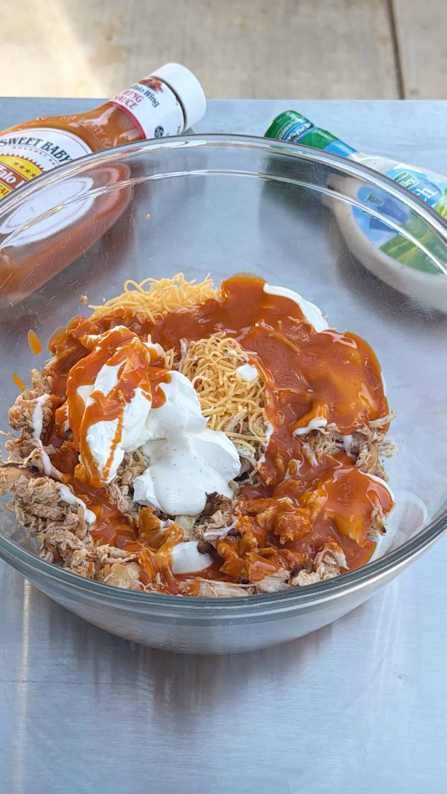 A glass bowl filled with shredded chicken, shredded cheese, ranch dressing, and buffalo sauce, with bottles of ranch and buffalo sauce in the background on a metal surface.  This is the filling for buffalo chicken sliders