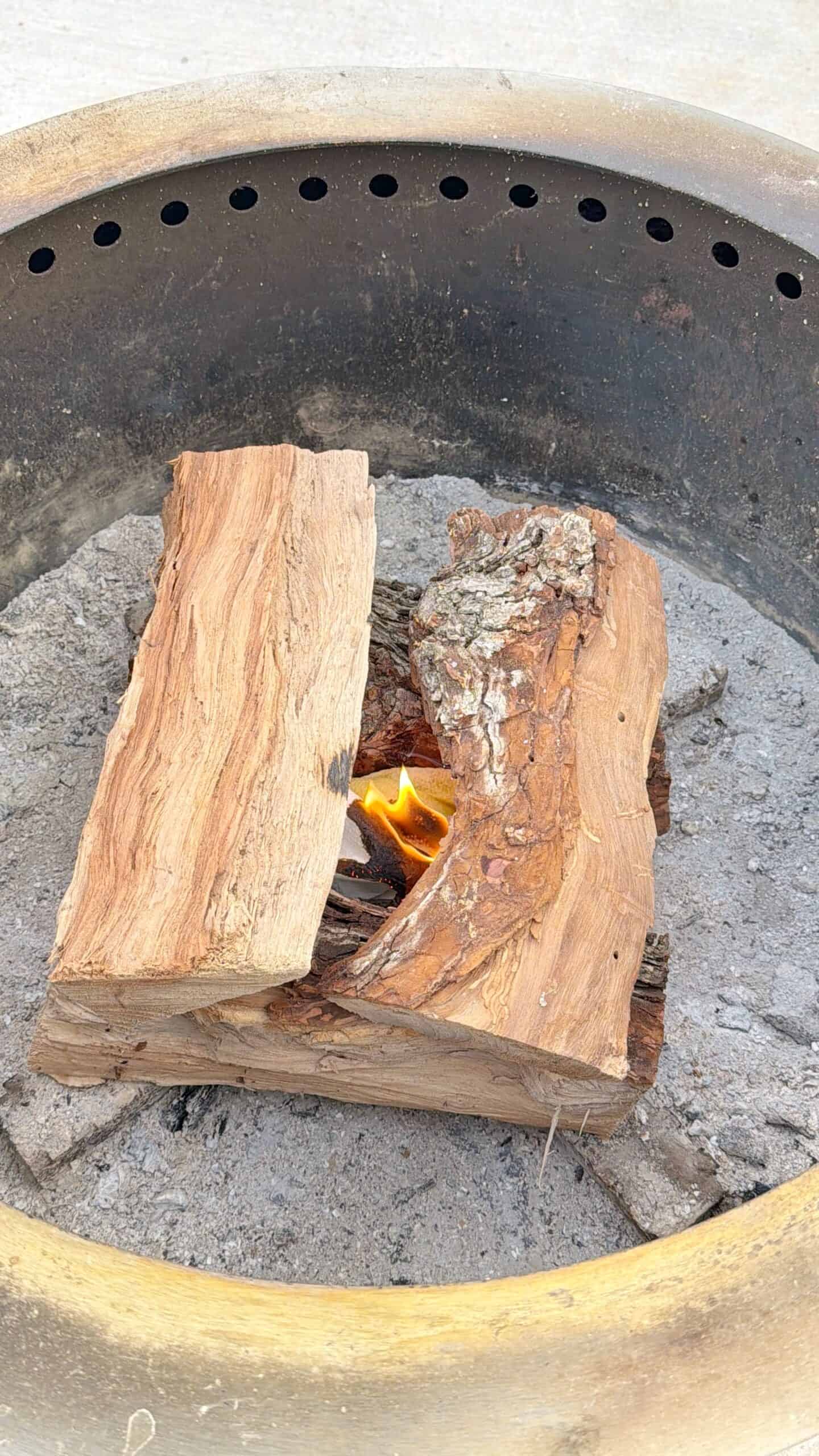 Two wooden logs placed inside a circular metal fire pit with a small flame burning between them, surrounded by ash and gray debris at the bottom of the pit.