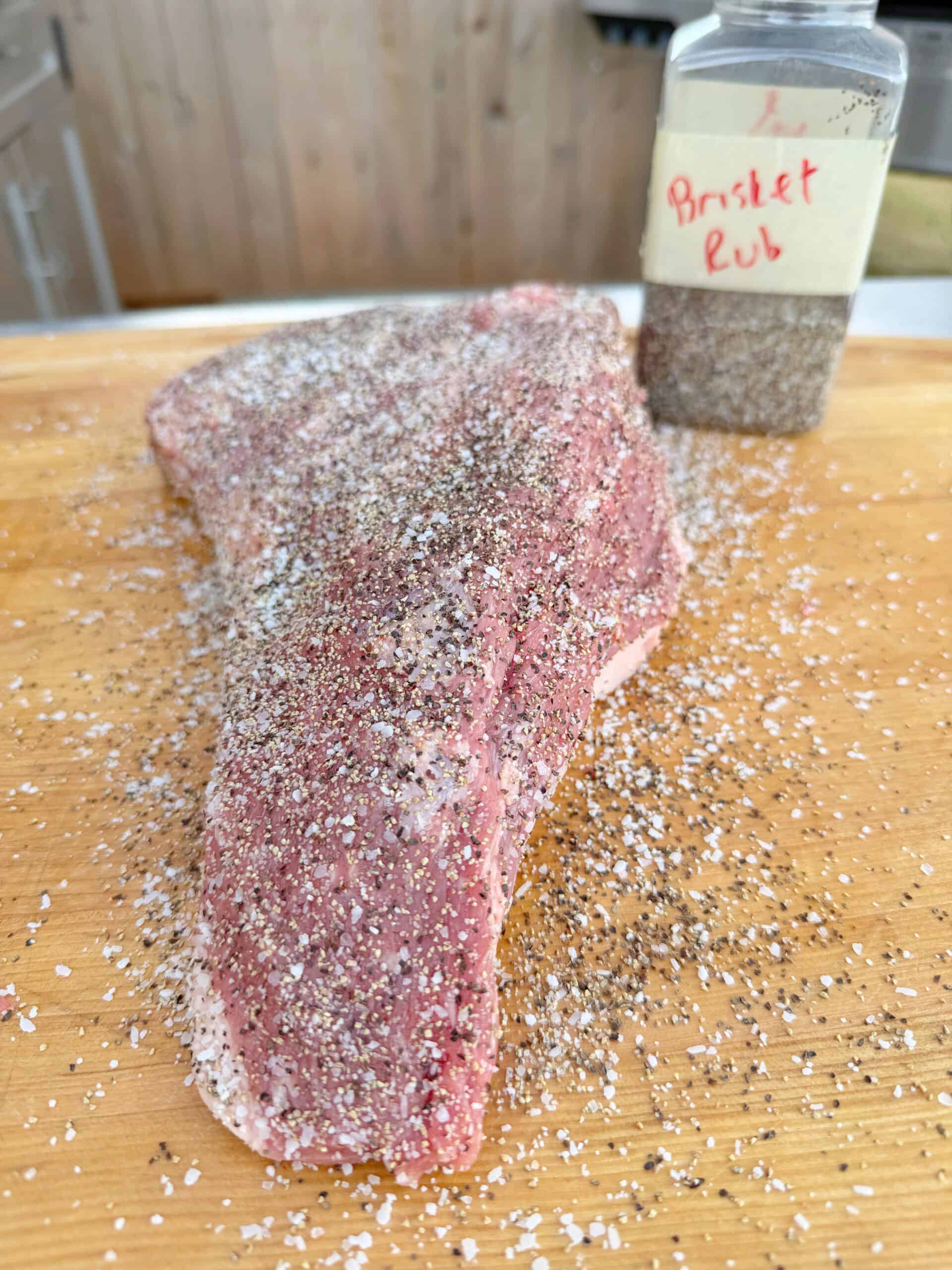 A raw brisket covered in coarse seasoning sits on a wooden cutting board next to a clear container labeled Brisket Rub, with seasoning scattered around.