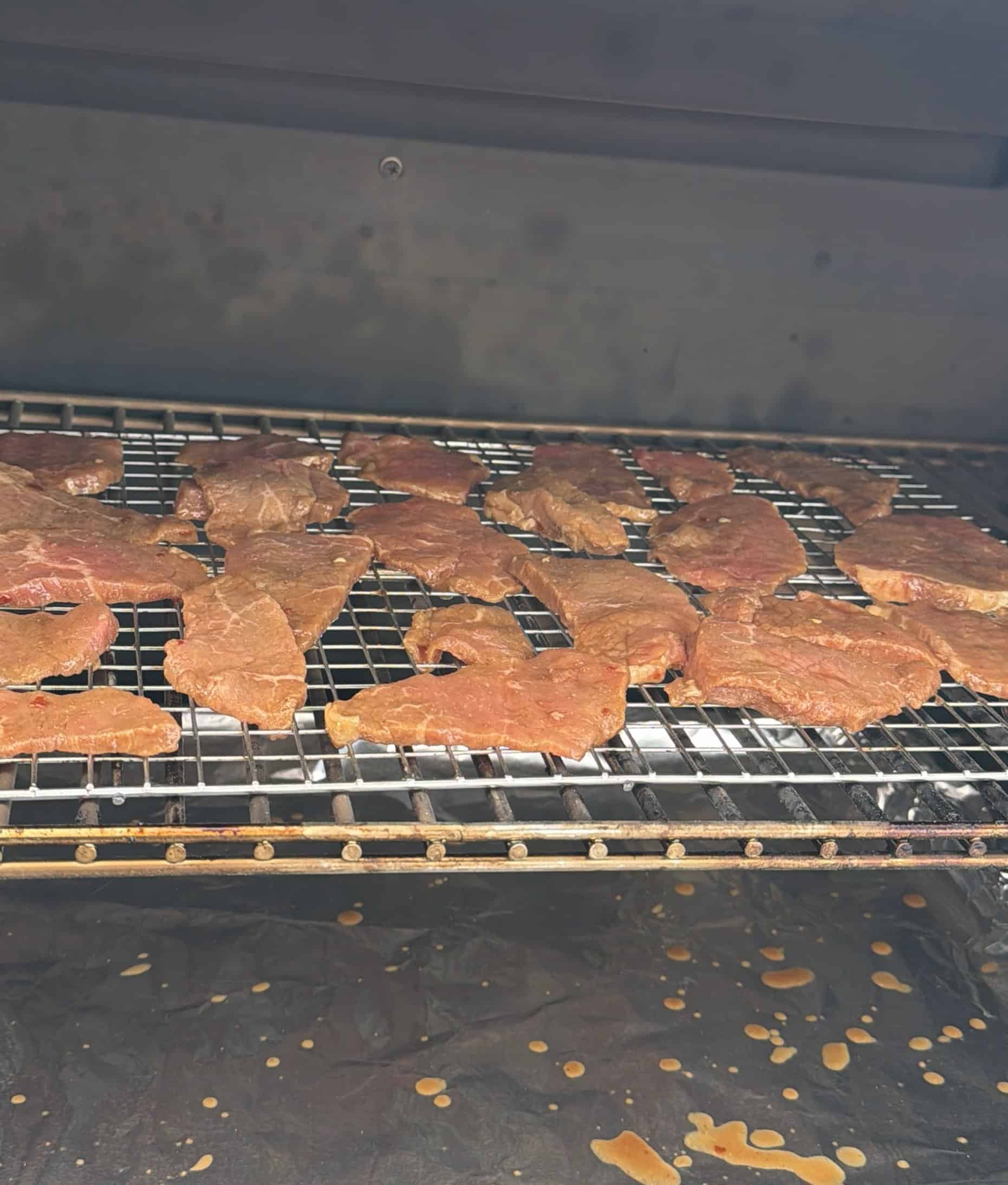 Sliced pieces of marinated meat are arranged on a wire rack inside a smoker or oven, in the process of being dried or cooked, with drippings visible on the tray below.  Food cooking is jalapeno teriyaki beef jerky