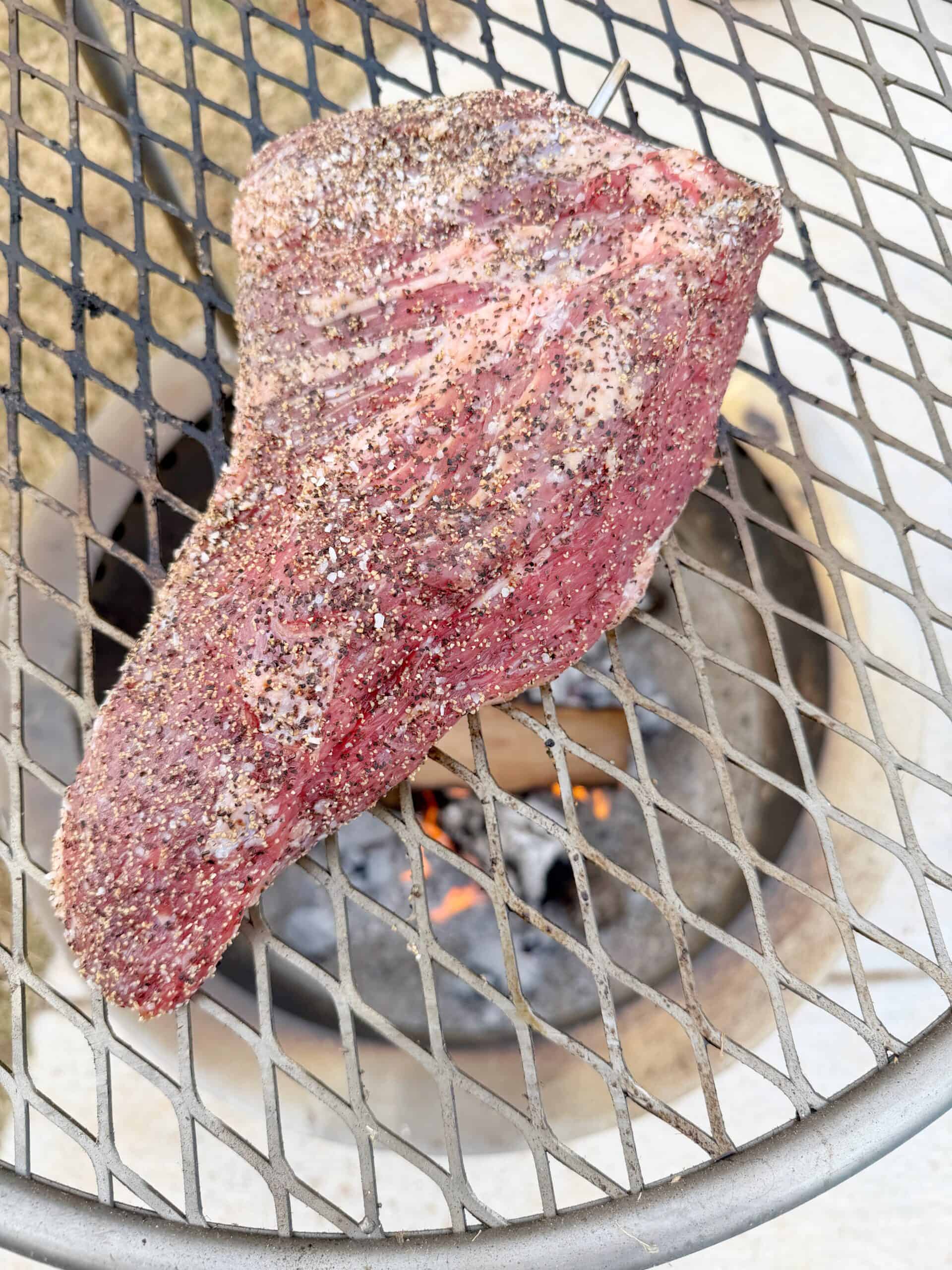 A seasoned, raw steak sits on a grill grate above a wood fire, with flames and burning logs visible below.