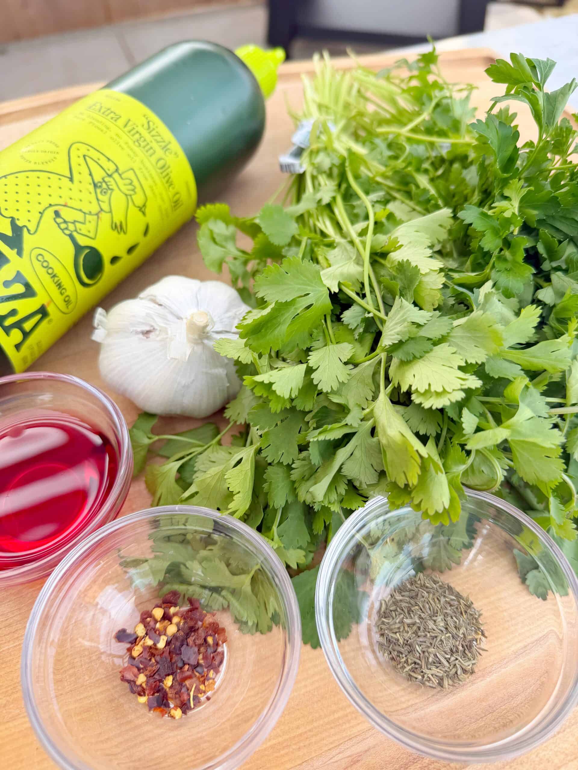 Fresh parsley, cilantro, a garlic bulb, olive oil bottle, red wine vinegar in a small bowl, dried chili flakes, and dried oregano on a wooden surface, all ingredients for preparing Smoked Chimichurri 