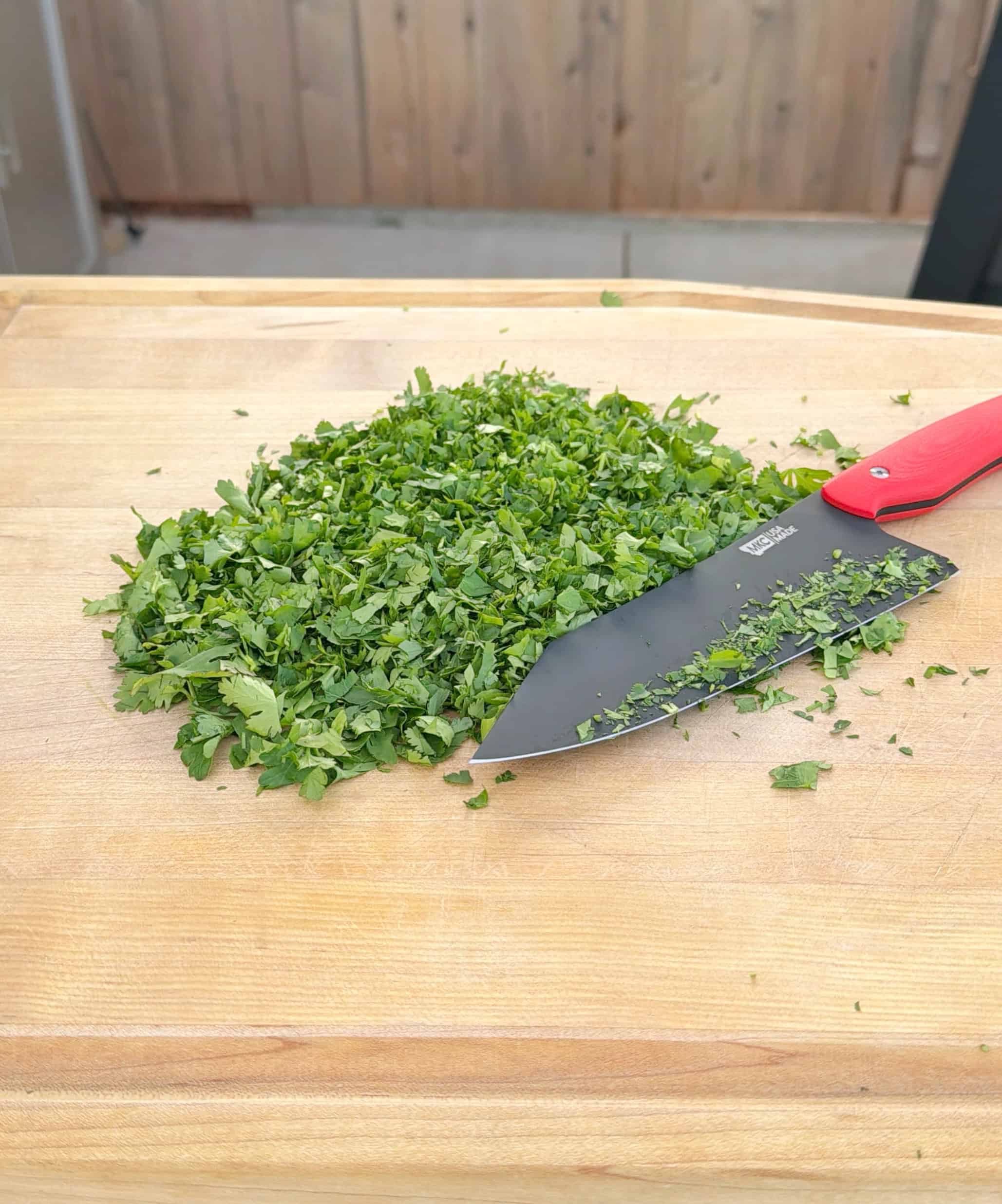 A pile of freshly chopped green herbs on a wooden cutting board, with a large black knife featuring a red handle resting next to the herbs.