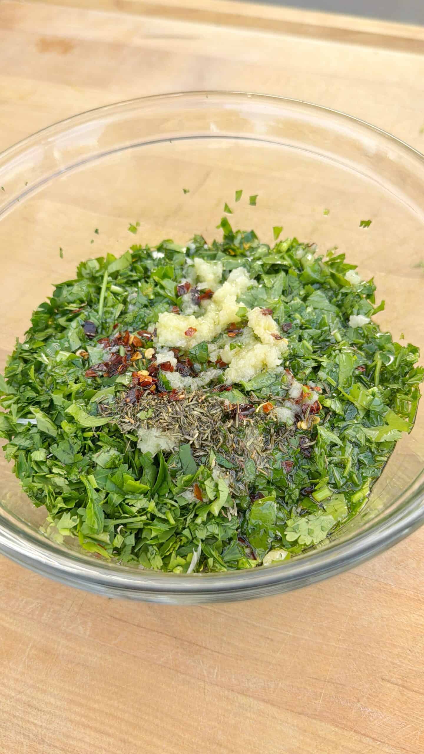 A glass bowl on a wooden surface filled with chopped fresh herbs, minced garlic, dried chili flakes, and other seasonings ready to be mixed.