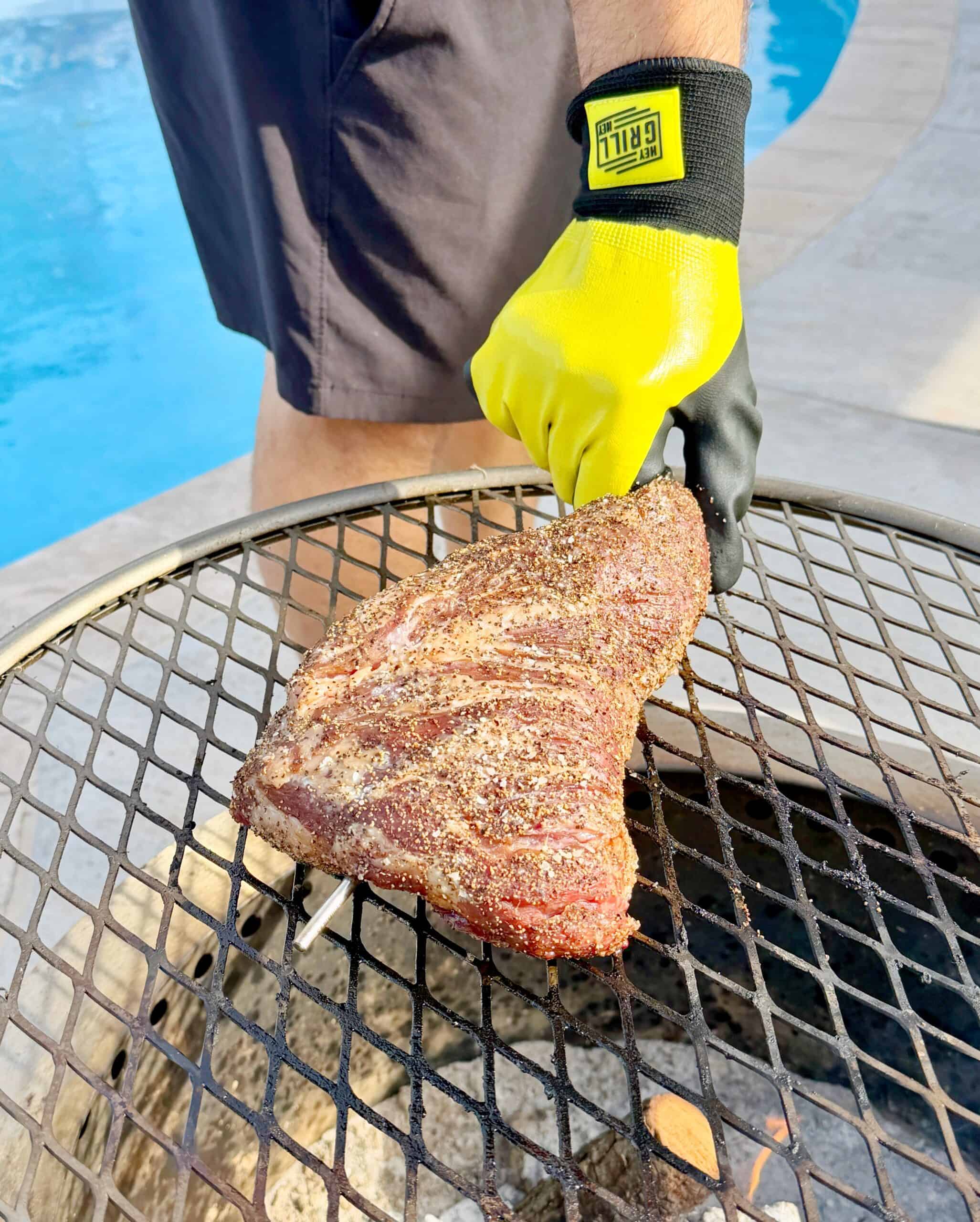 A person wearing yellow and black grilling gloves is placing a seasoned cut of meat on a grill over a fire, with a swimming pool and patio visible in the background.