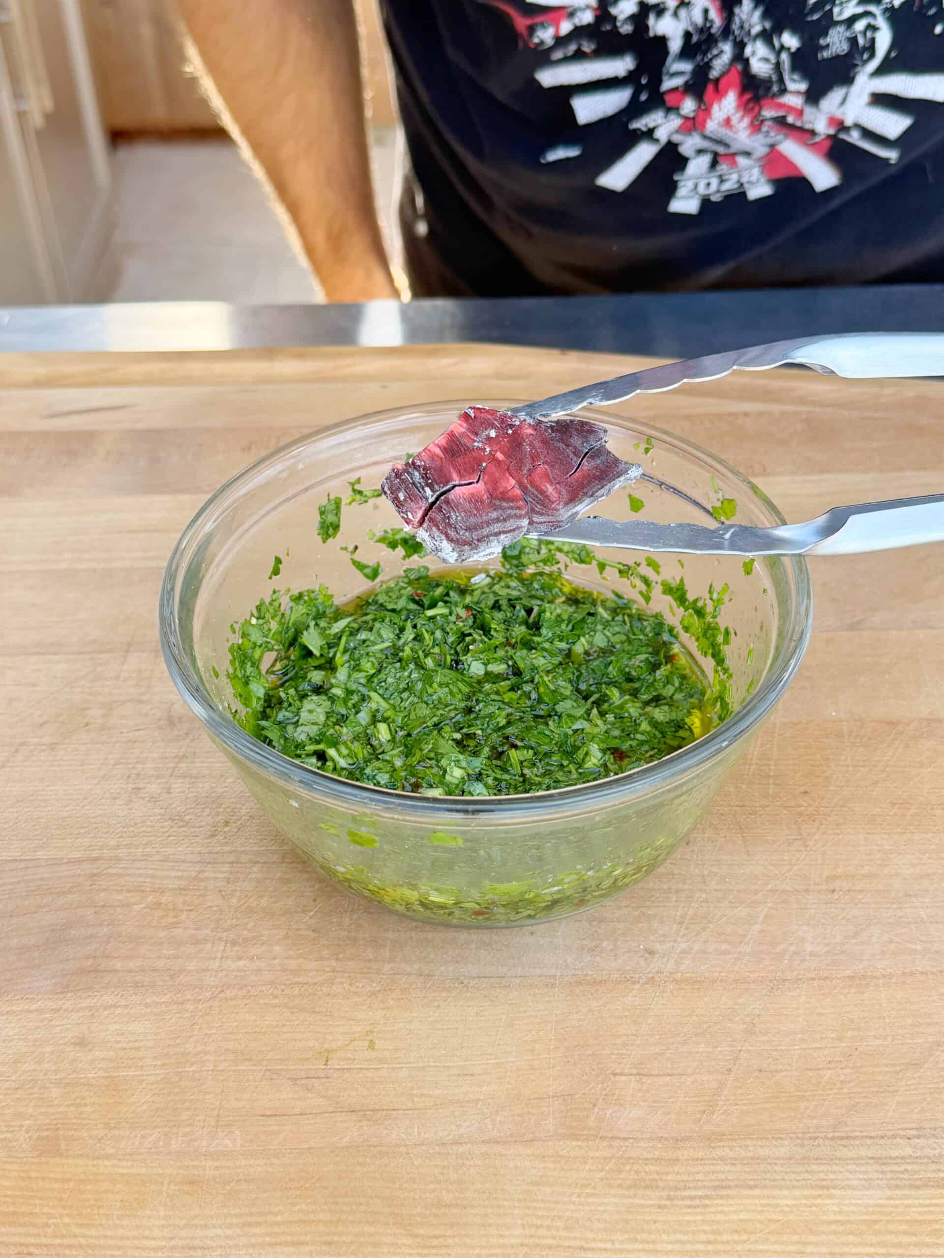 A person holding a piece of raw steak with metal tongs above a glass bowl filled with chopped green herbs on a wooden surface.