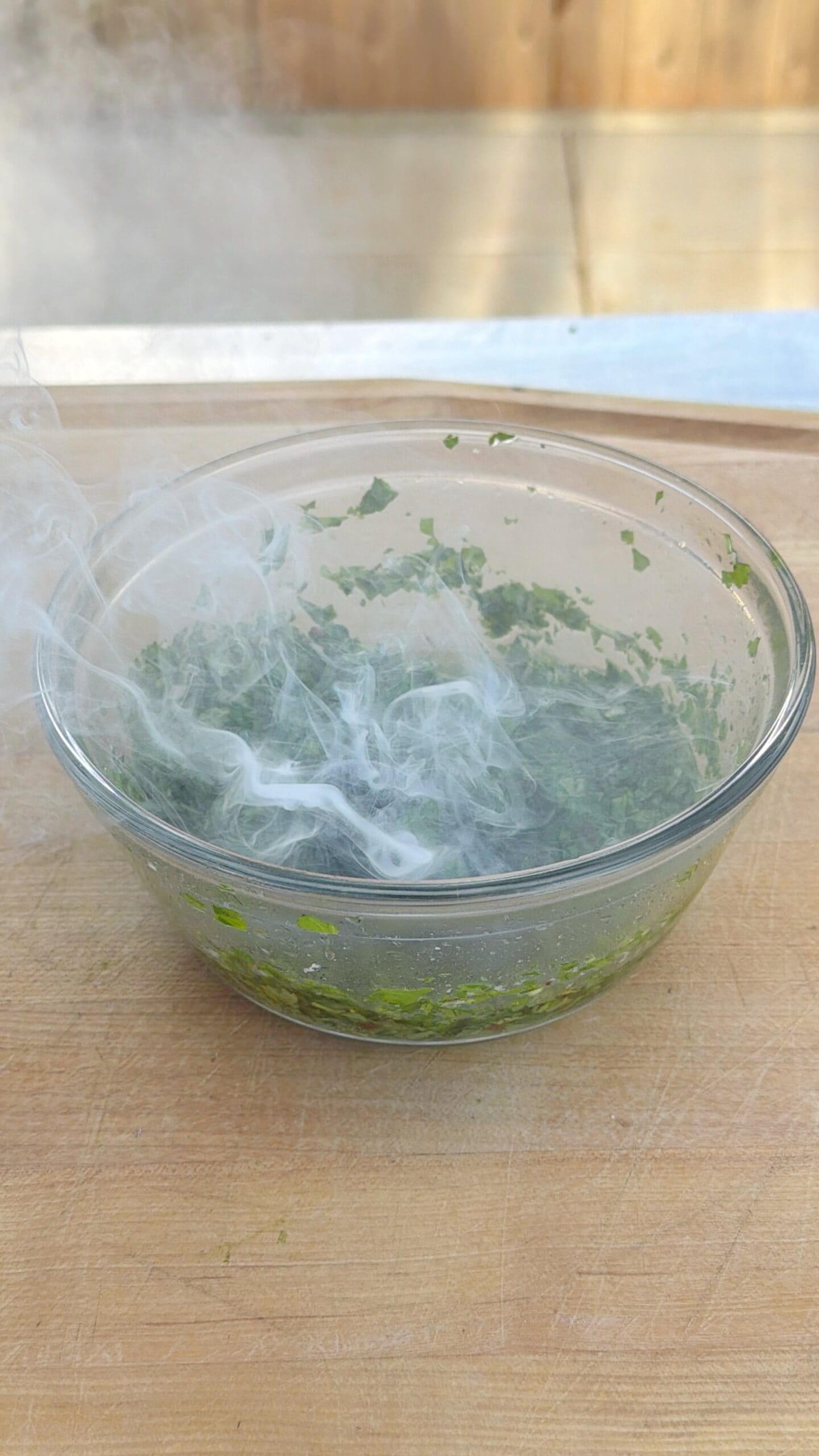A glass bowl with chopped green herbs sits on a wooden surface, with wisps of white smoke swirling above the mixture.