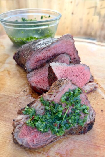 Sliced medium-rare steak on a wooden cutting board, topped with fresh green chimichurri sauce, with a glass bowl of extra sauce in the background.