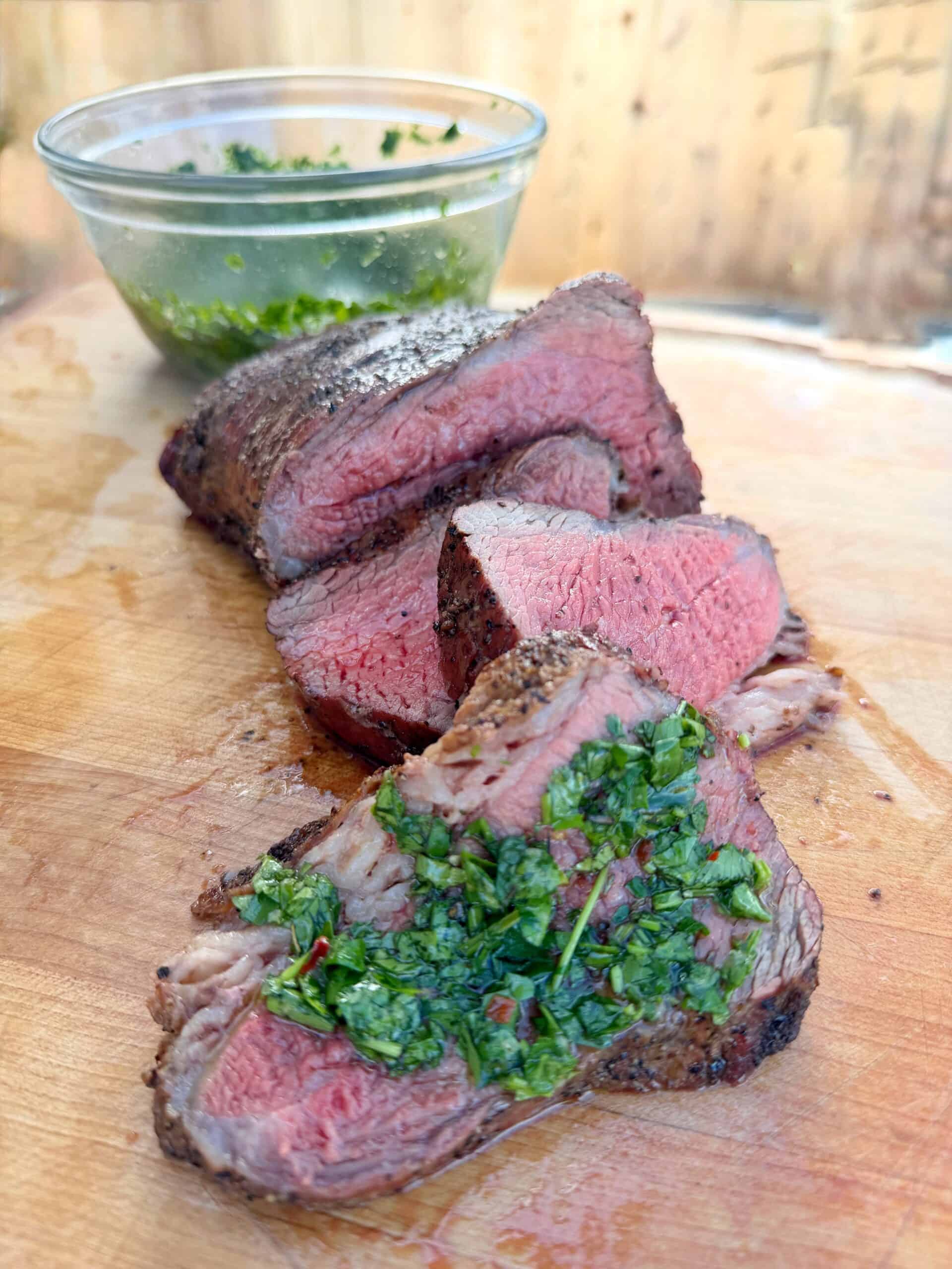 Sliced medium-rare steak on a wooden cutting board, topped with fresh green chimichurri sauce, with a glass bowl of extra sauce in the background.