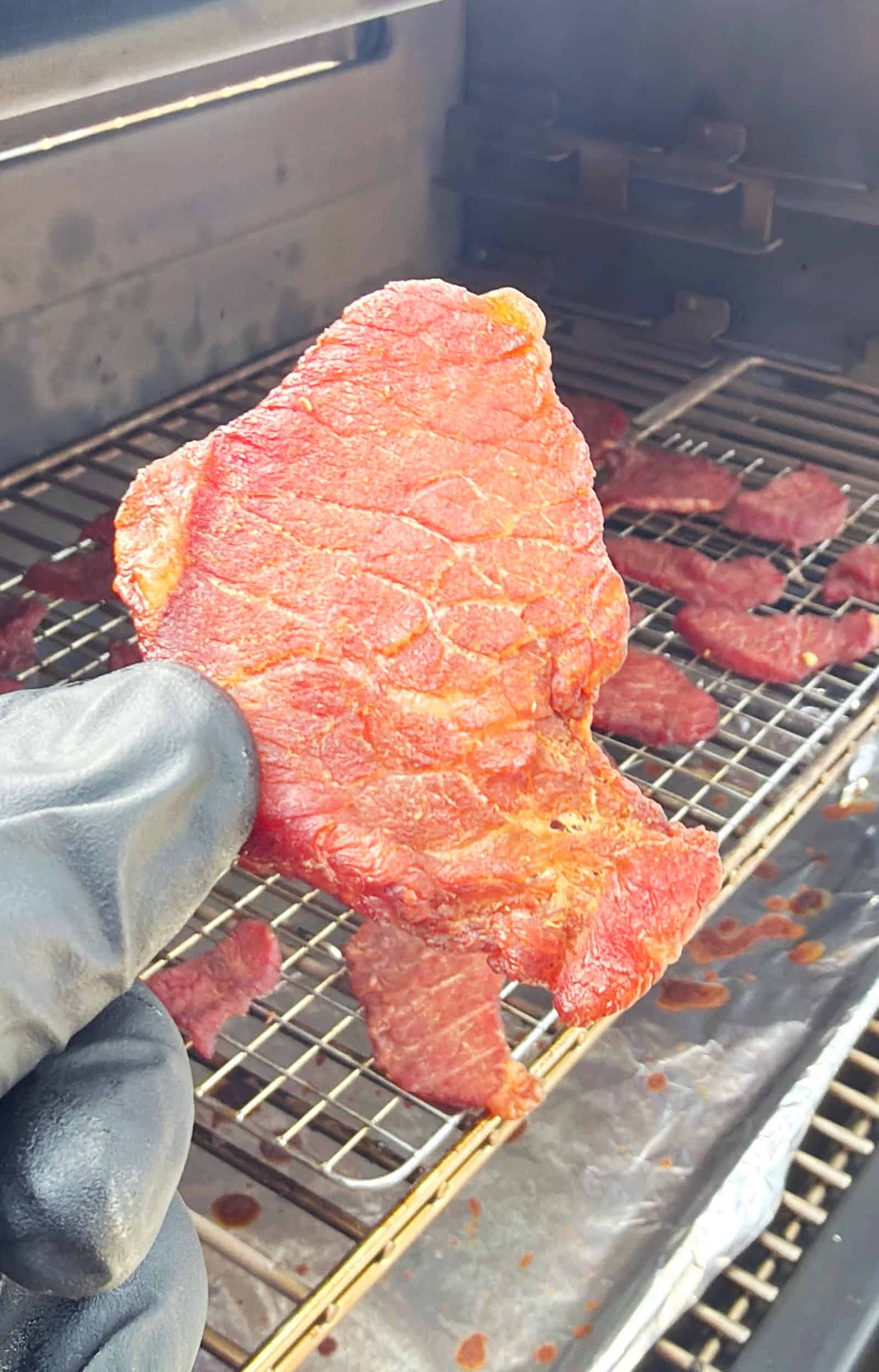 A gloved hand holds up a piece of seasoned raw beef above a grill rack, with several other strips of beef laying flat on the wire rack inside a smoker or grill.