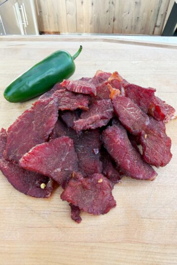 A pile of sliced beef jerky sits on a wooden cutting board with a whole green jalapeño pepper placed beside it. The background features part of an outdoor kitchen and a wooden fence.