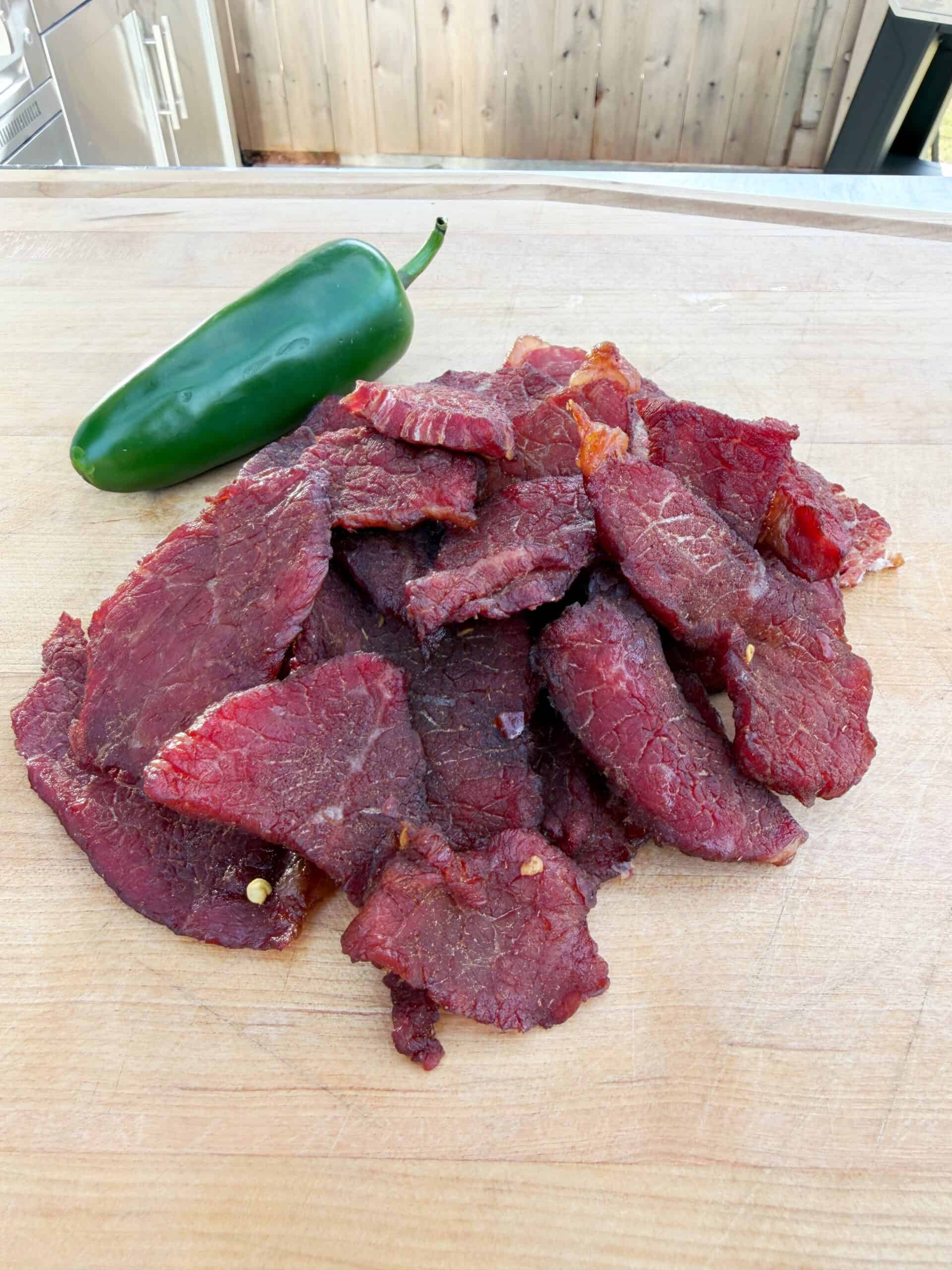 A pile of sliced beef jerky sits on a wooden cutting board with a whole green jalapeño pepper placed beside it. The background features part of an outdoor kitchen and a wooden fence.