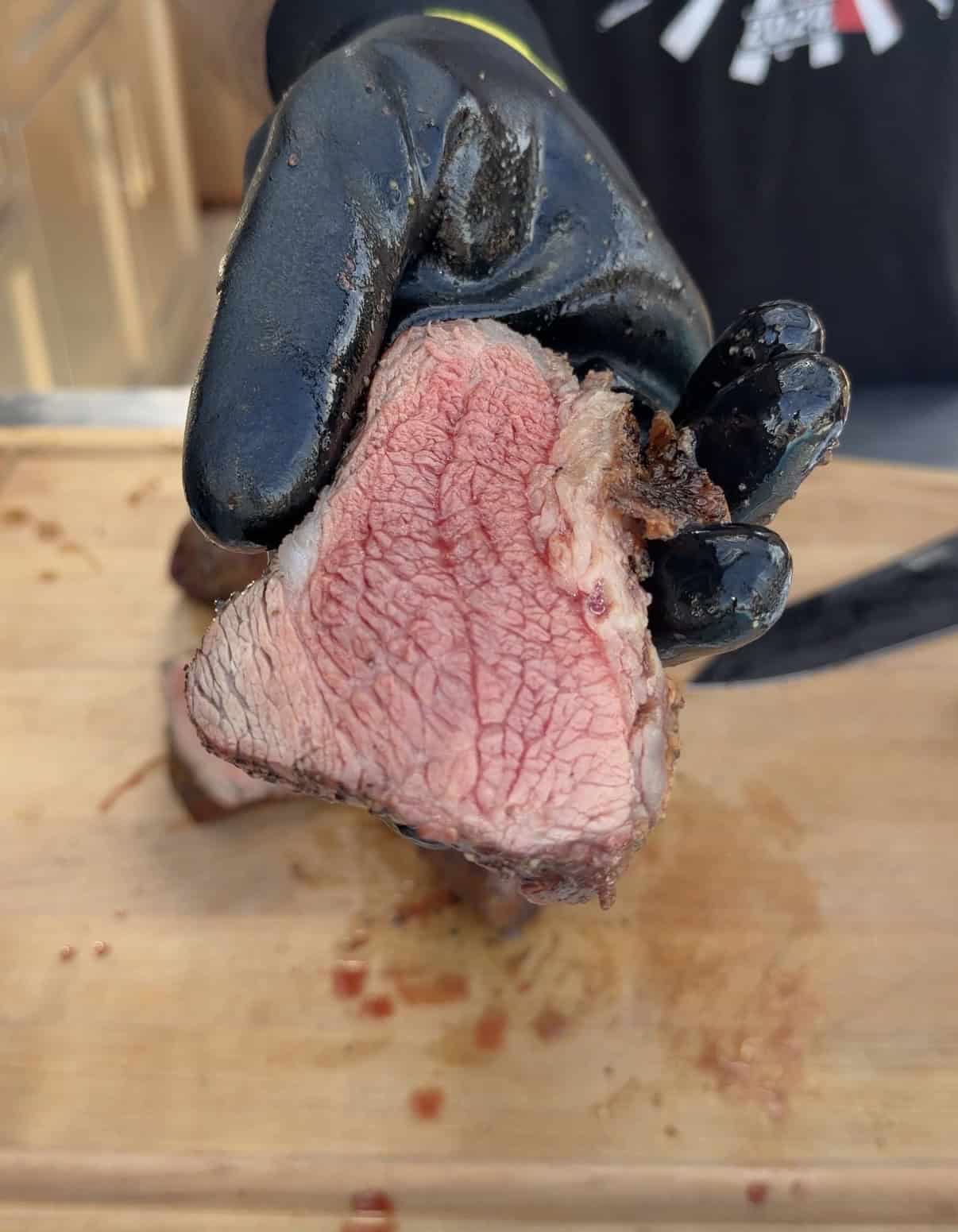 A person wearing a black glove holds up a juicy, medium-rare slice of beef over a wooden cutting board, showing the pink interior and seared outer edge.
