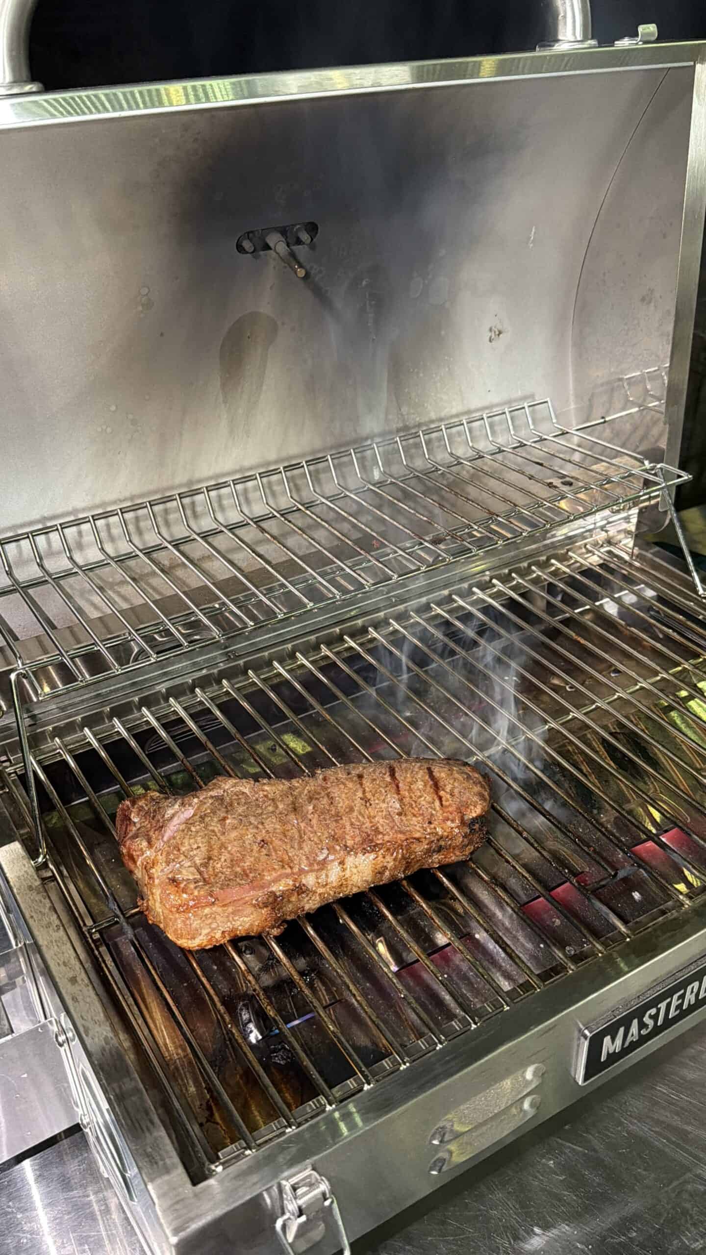 A thick, seasoned steak is grilling on the lower rack of an open stainless steel Masterbuilt grill, with smoke rising and grill marks visible, while the upper rack is empty.