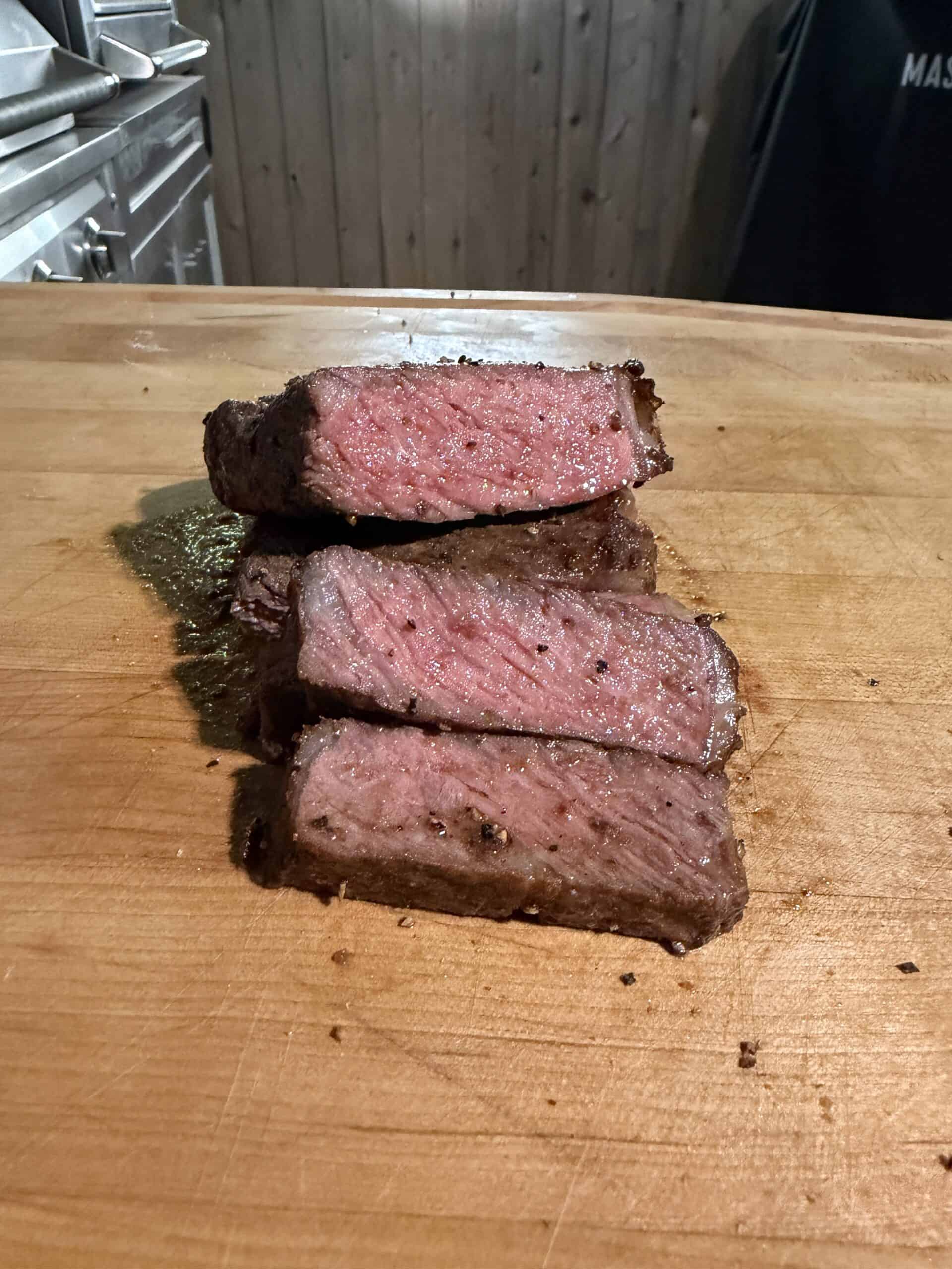 Three slices of medium-rare steak are stacked on a wooden cutting board, showing a pink, juicy interior and seared, seasoned exterior. The background includes a wood-paneled wall and part of a kitchen.