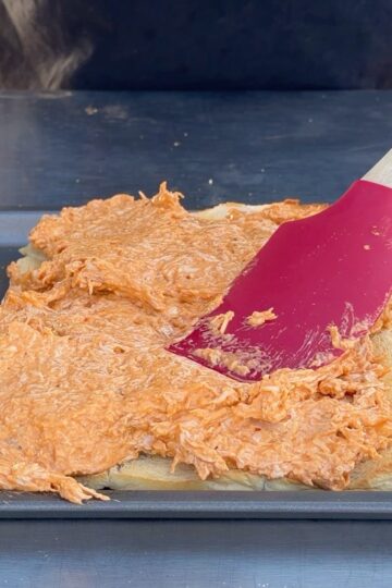 A person spreads a creamy, orange shredded chicken mixture onto a sheet of dough in a baking pan using a red spatula.