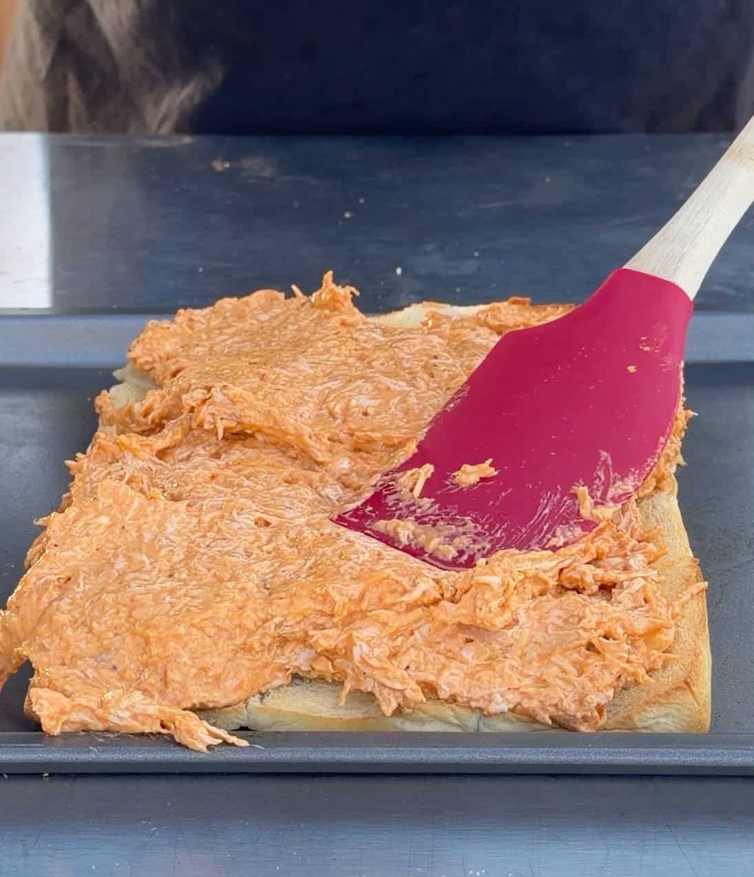 A person spreads a creamy, orange shredded chicken mixture onto a sheet of dough in a baking pan using a red spatula.