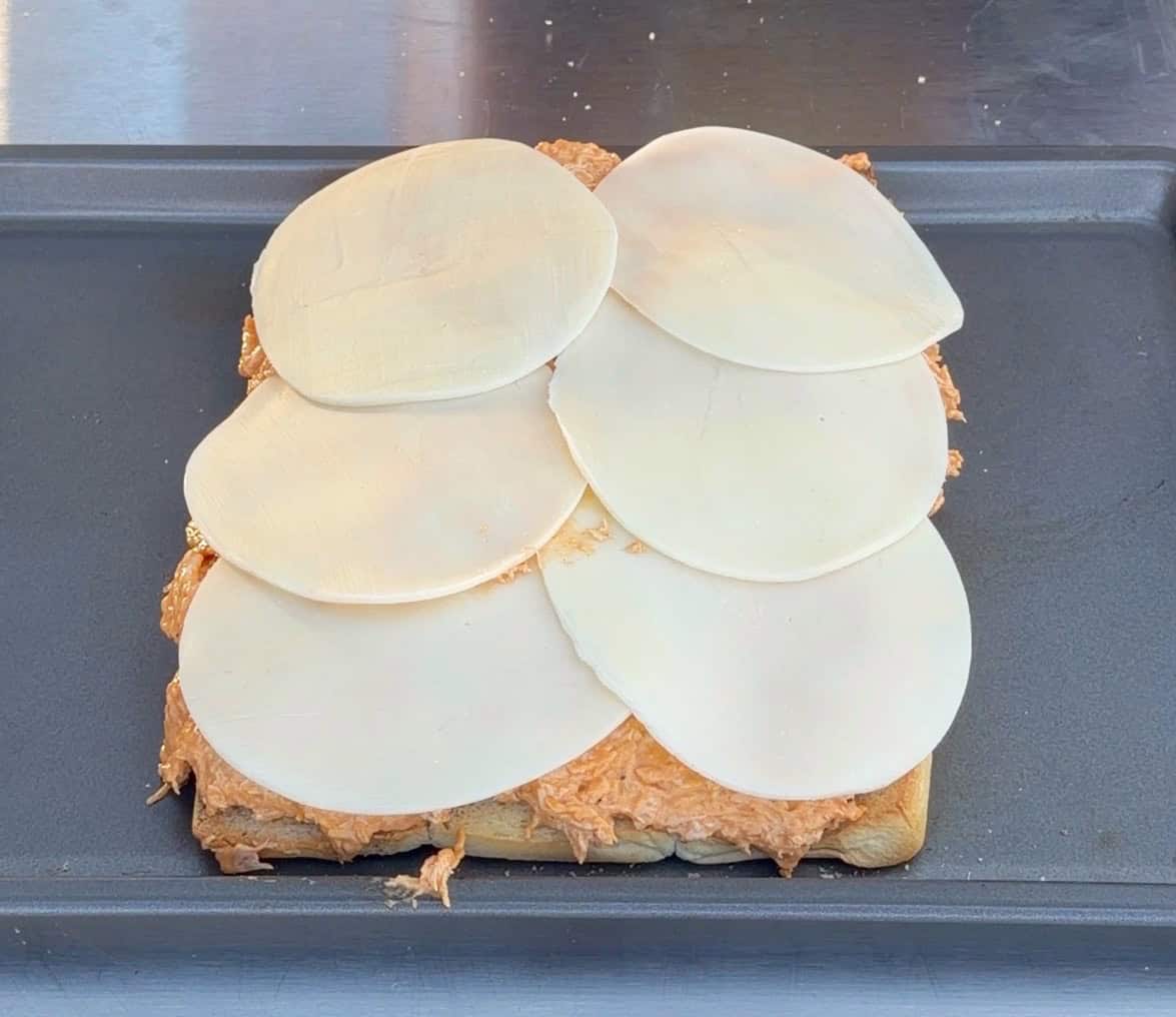 A baking tray holds an open sandwich topped with shredded chicken and six overlapping slices of white cheese.
