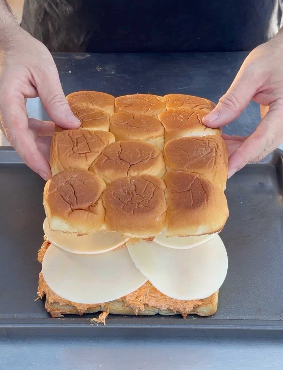 A person places the top layer of a pack of slider buns onto shredded chicken and provolone cheese on a baking tray, preparing a batch of sliders.