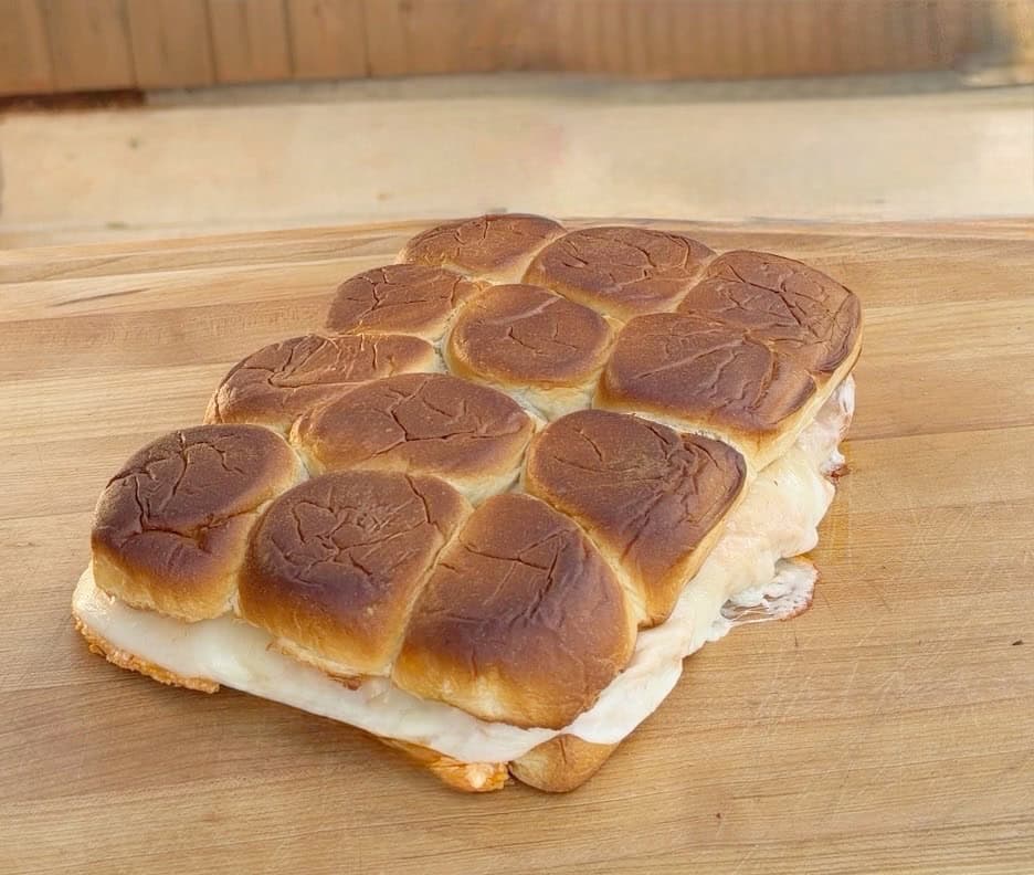A rectangular tray of baked slider sandwiches with golden-brown tops and melted cheese oozing from the sides, placed on a wooden surface.